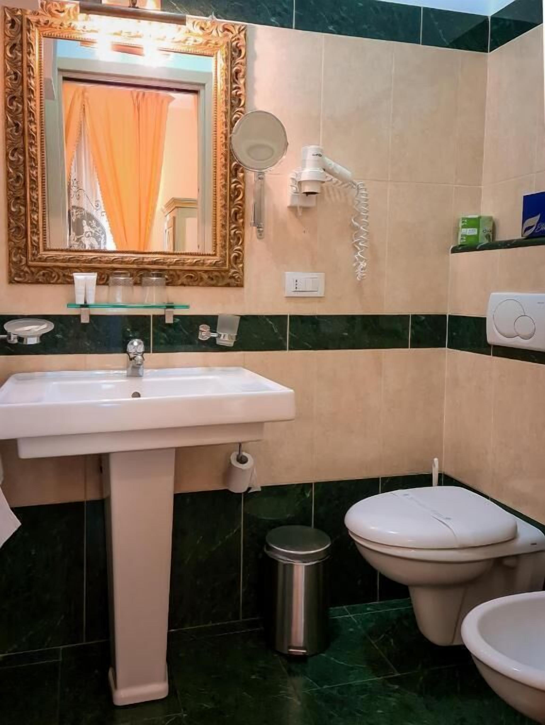 A bathroom with a white sink, a toilet, a trash bin, a mirror with a decorative frame, and a towel rack.