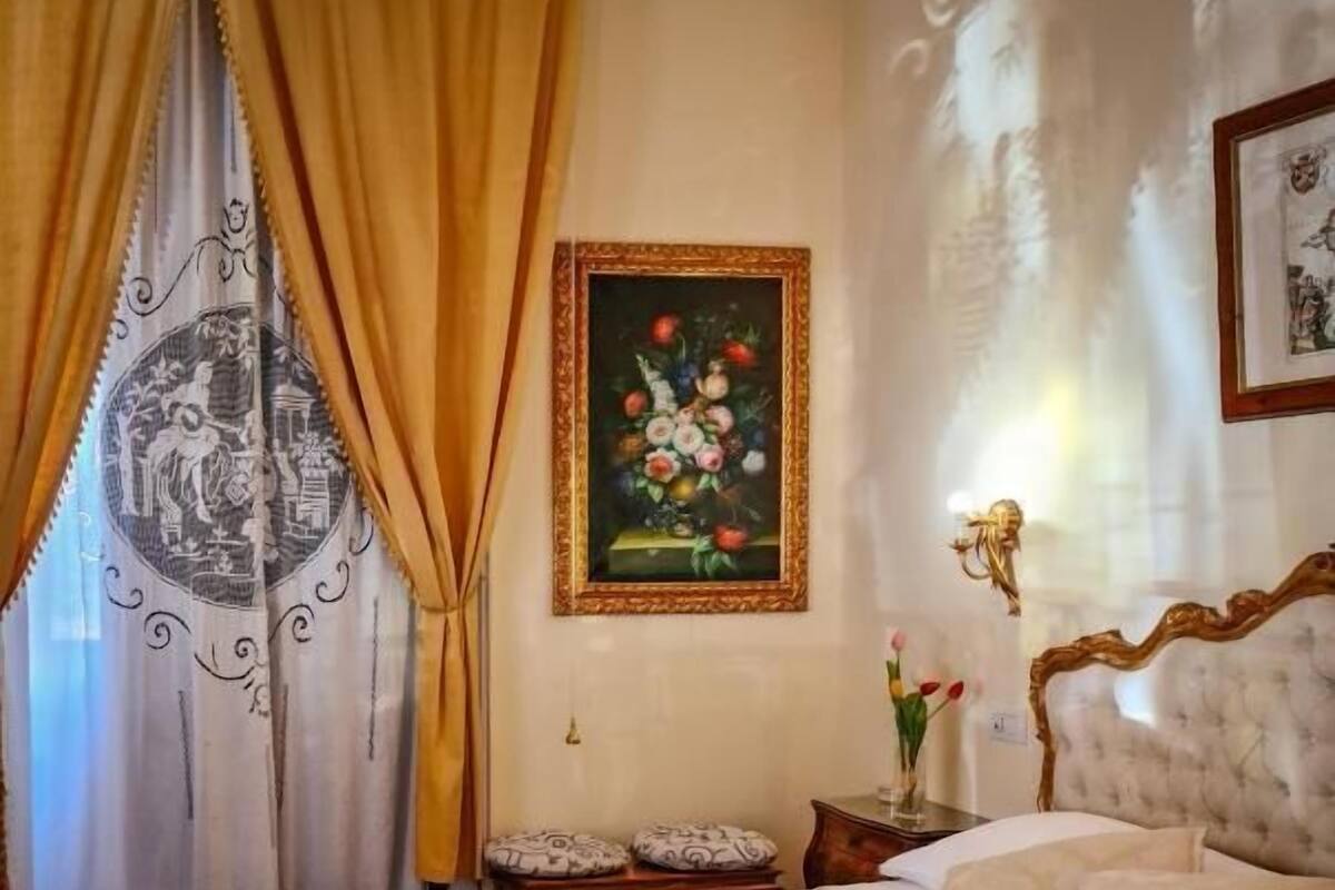 Een slaapkamer met een groot bed, een nachtkastje, een lijst met bloemen en gordijnen.