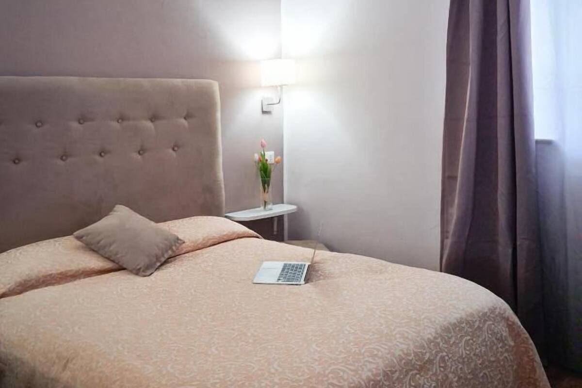 Een net opgemaakt bed met een laptop, een bedlampje en een vaas met bloemen.
