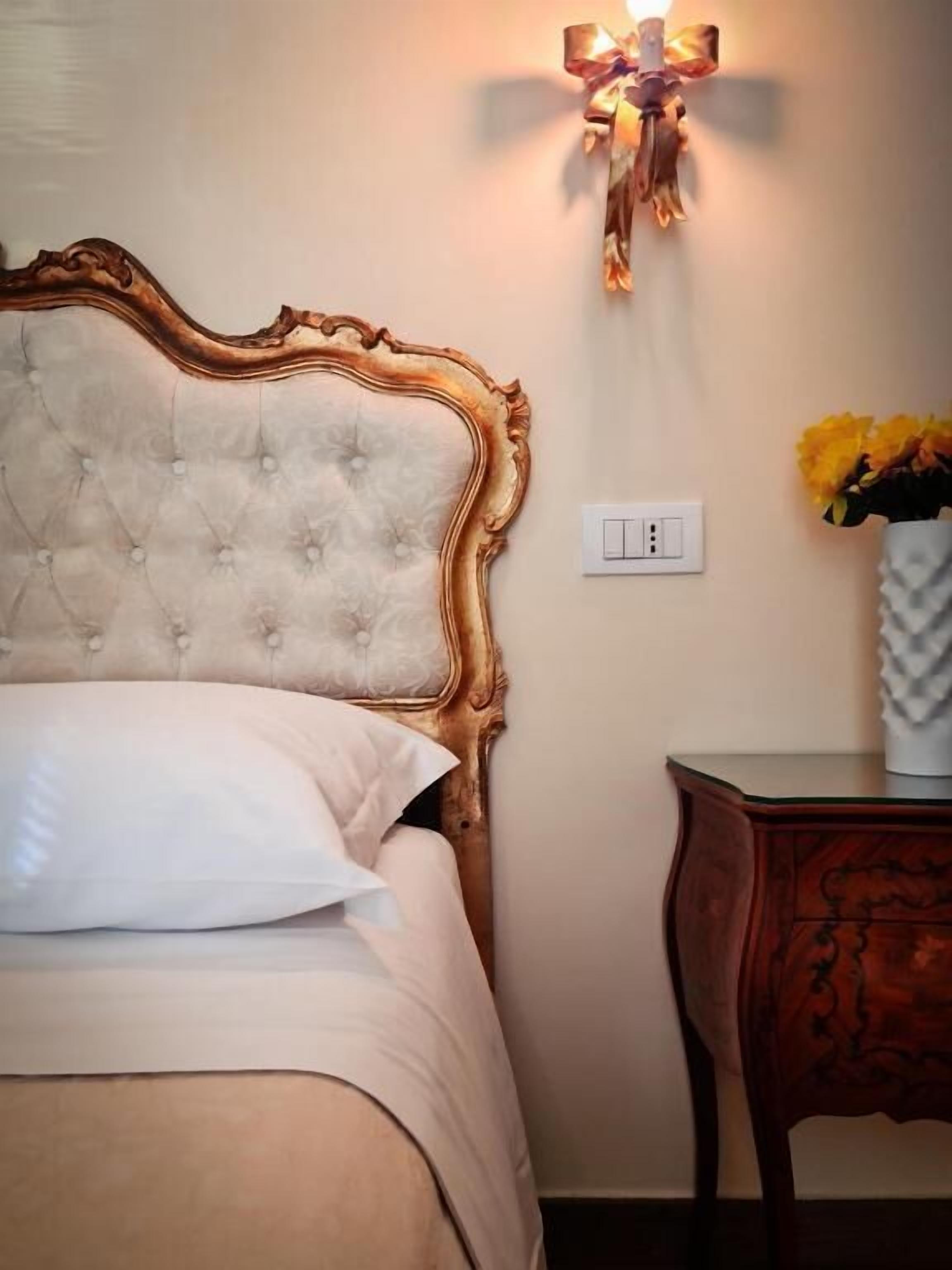 A bed with a tufted headboard, a bedside table with a vase of flowers, and a wall-mounted light fixture.