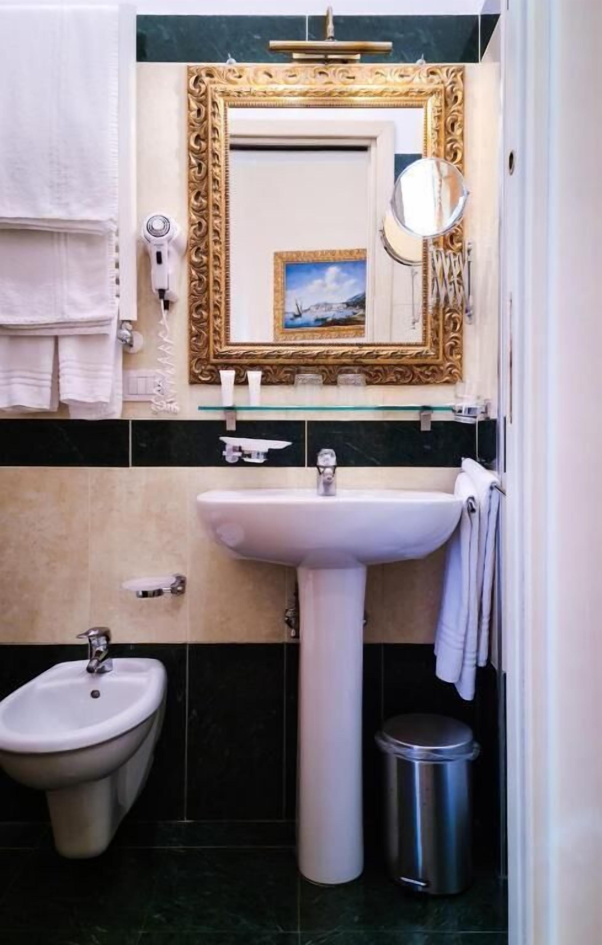 Une salle de bain équipée d’un lavabo sur colonne, d’un bidet, d’une poubelle et d’un miroir décoratif doté d’un cadre doré.