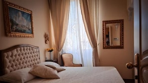 Ein ordentlich bezogenes Bett mit Kopfteil, ein Sessel, ein Gemälde, ein Spiegel und eine Tür mit Griff.