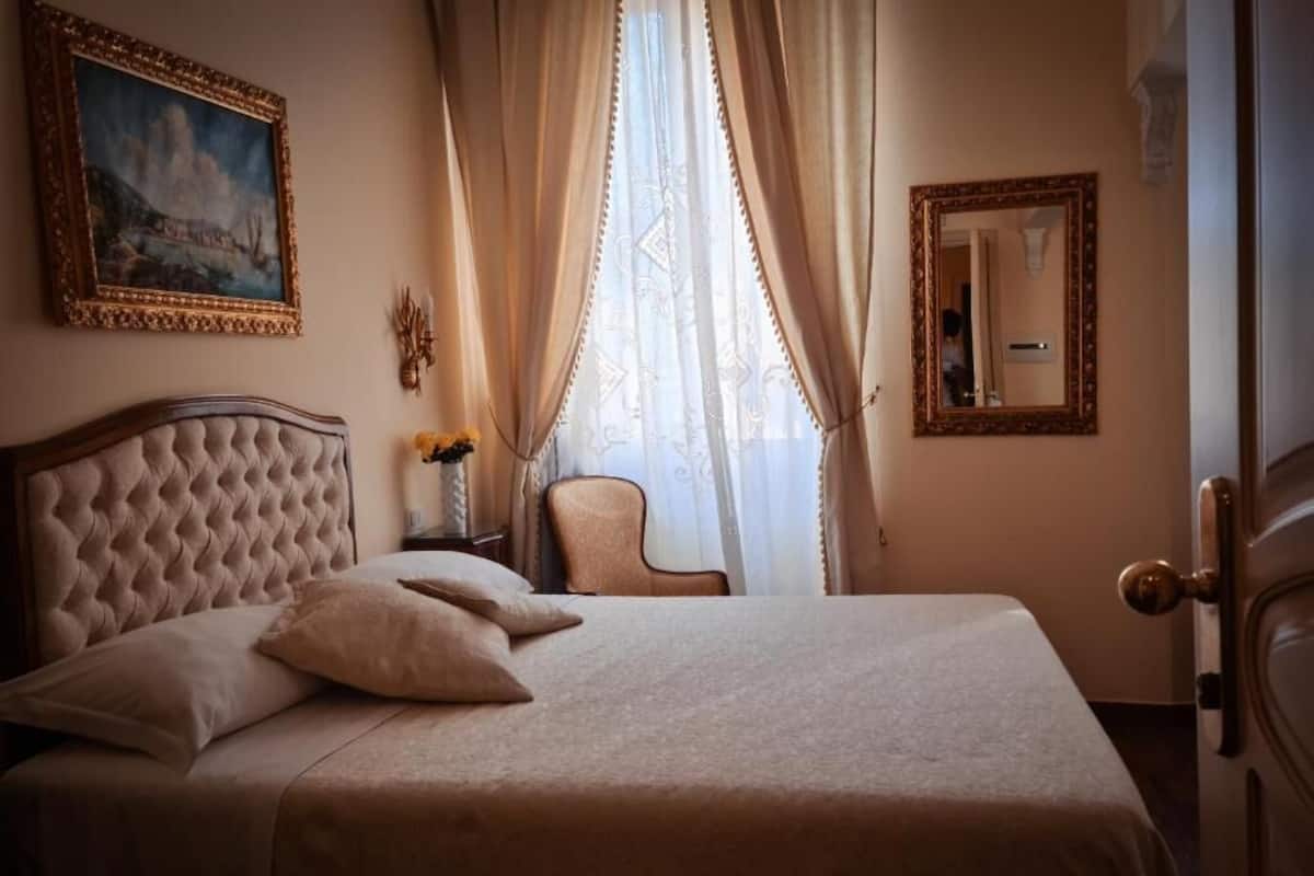 Een net opgemaakt bed met hoofdbord, een stoel, een schilderij, een spiegel en een deur met hendel.