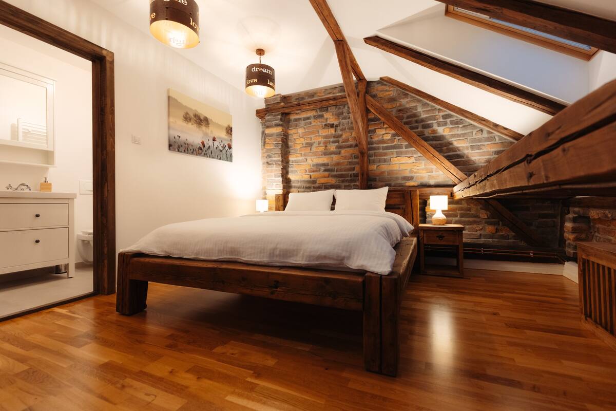 A bedroom with a wooden bed, a nightstand, a lamp, a painting, and a sloped ceiling with exposed beams.