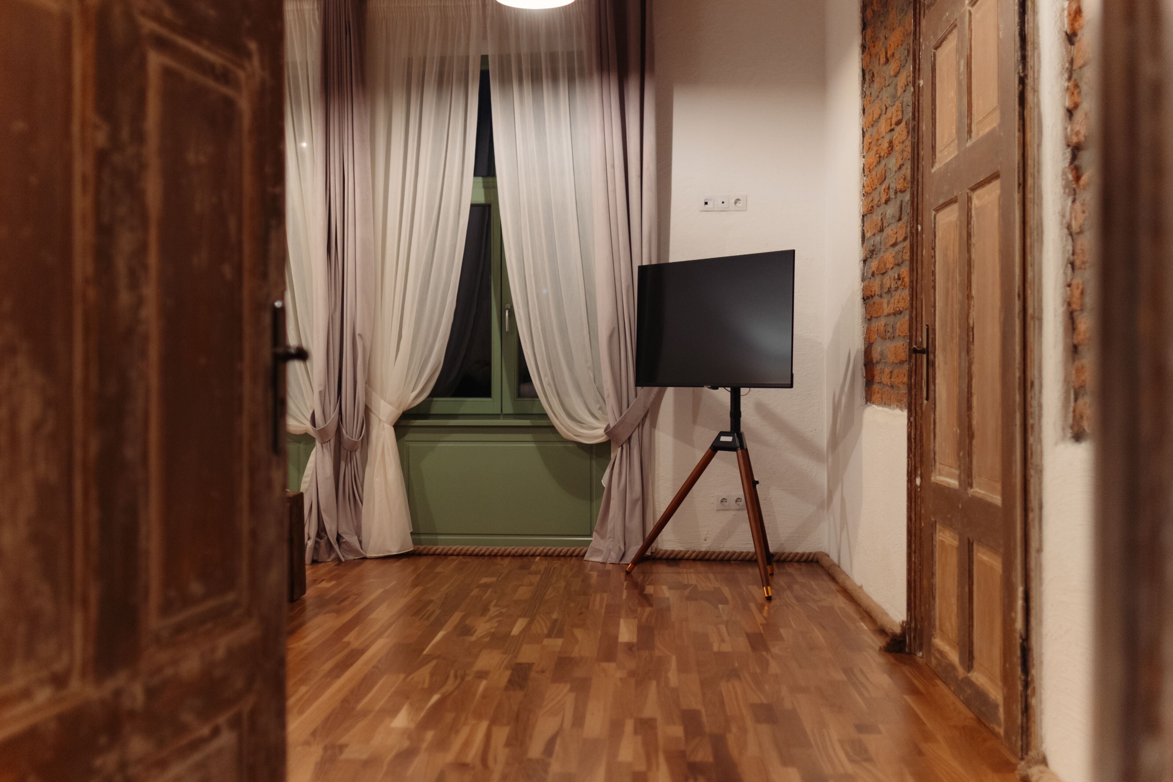 Une pièce avec un plancher en bois, une fenêtre avec des rideaux, un écran fixé sur un trépied et un mur en briques.