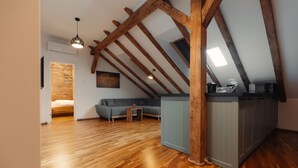 Une pièce de style « loft » avec des poutres apparentes, un plancher en bois, un canapé gris, une petite table et un coin cuisine.