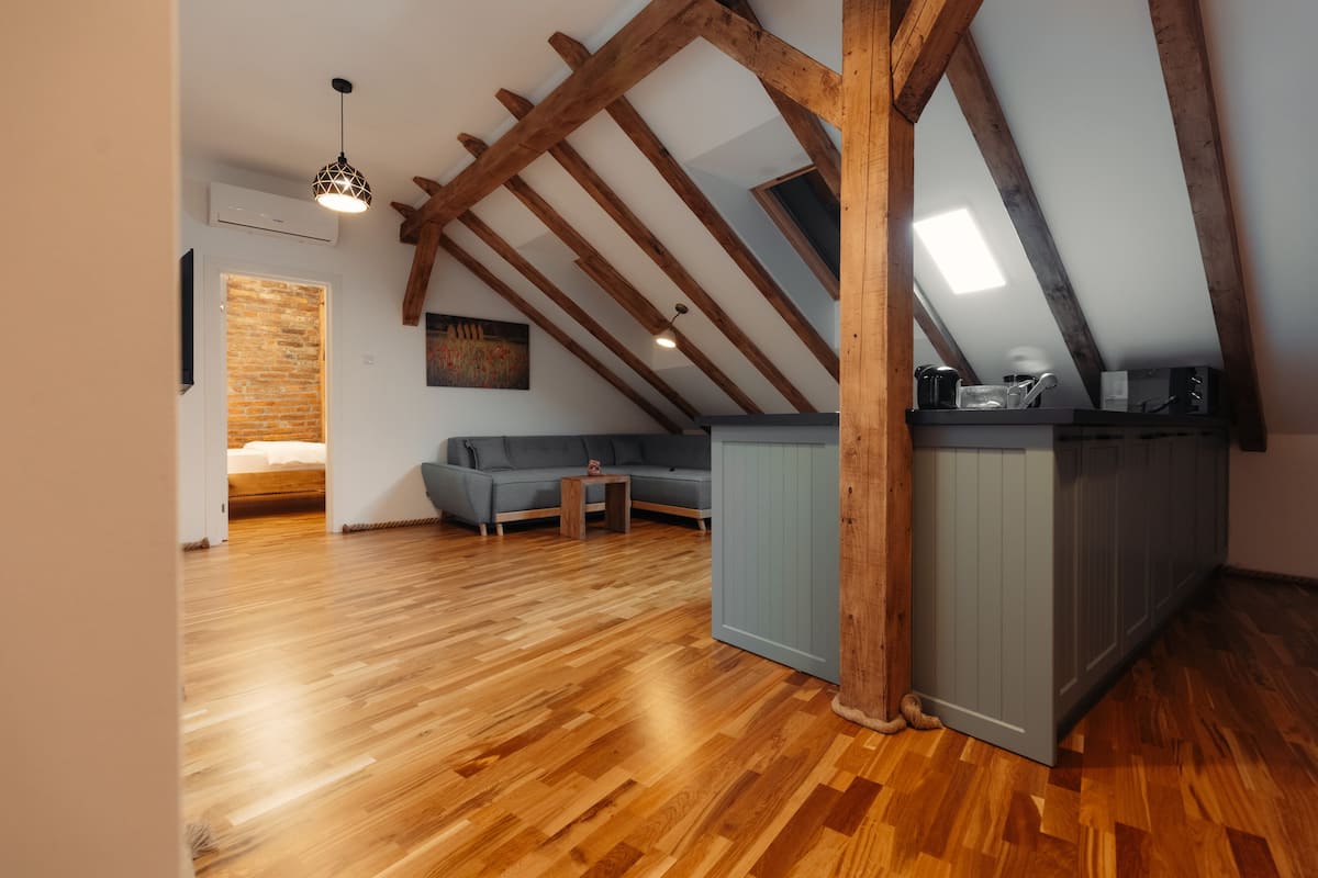 A loft-style room with exposed wooden beams, a wooden floor, a gray sofa, a small table, and a kitchenette area.