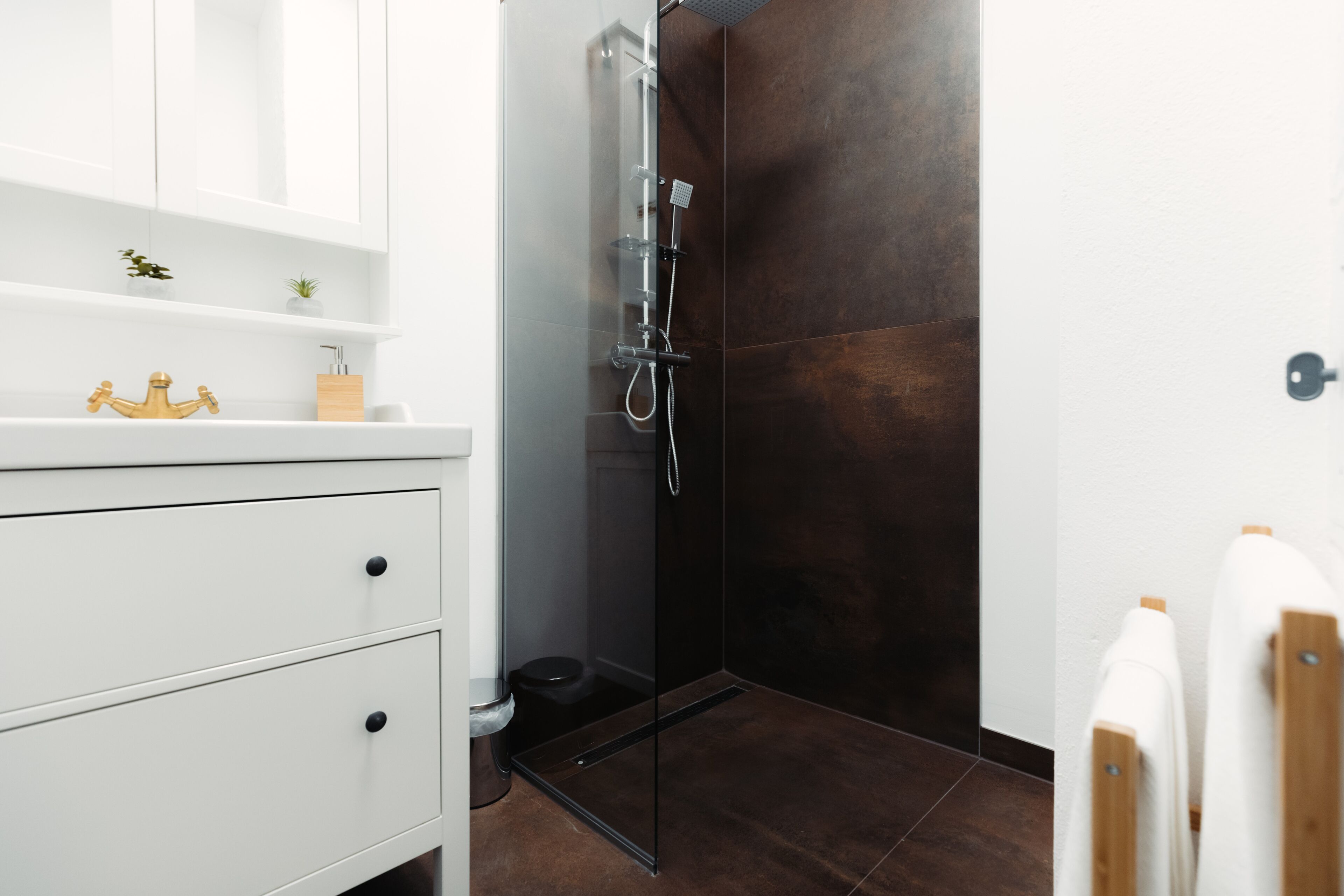 A modern bathroom with a white vanity, a glass shower enclosure, and dark brown tiles.