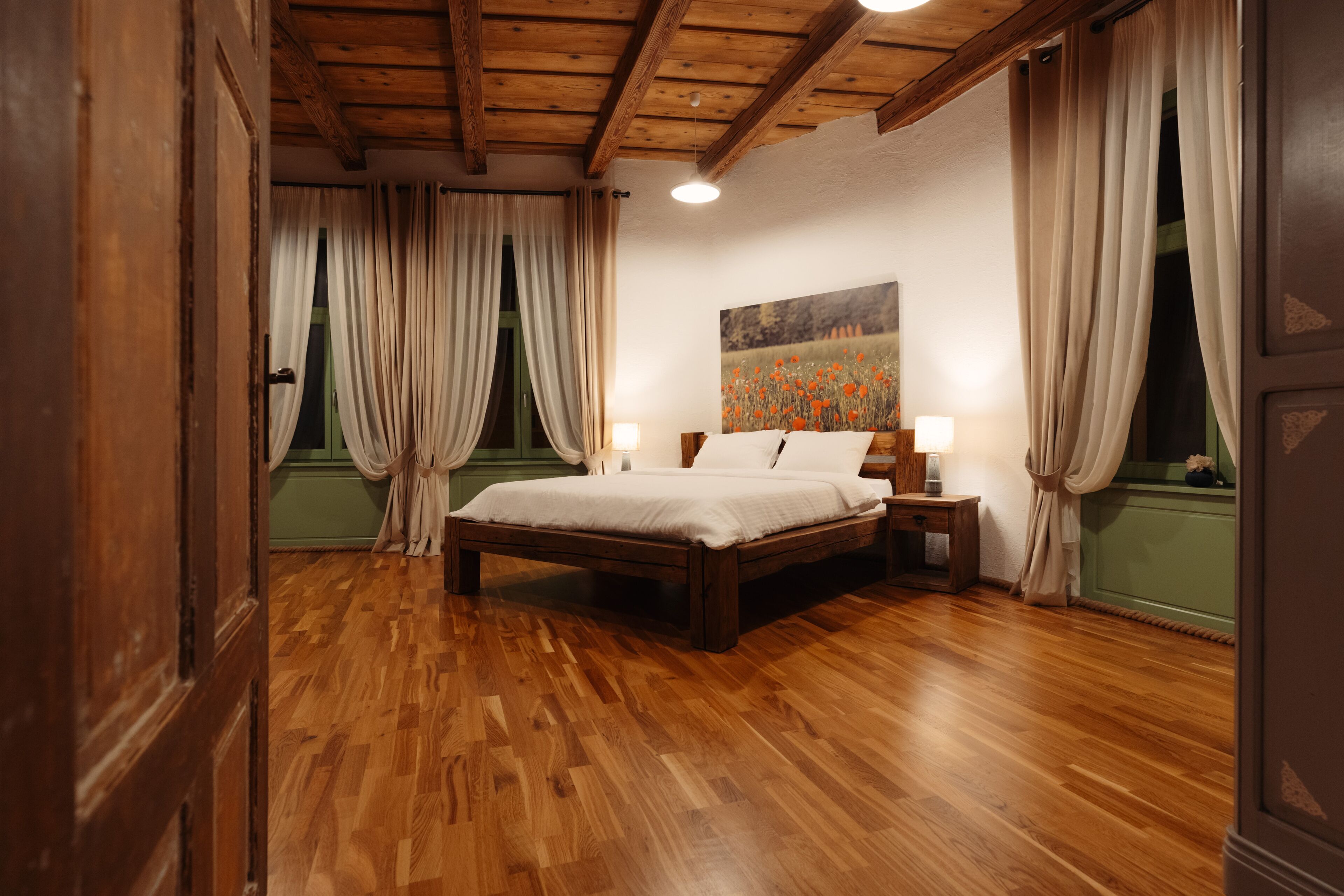 A bedroom with a wooden floor, a bed with white linens, a nightstand with a lamp, and a painting on the wall.