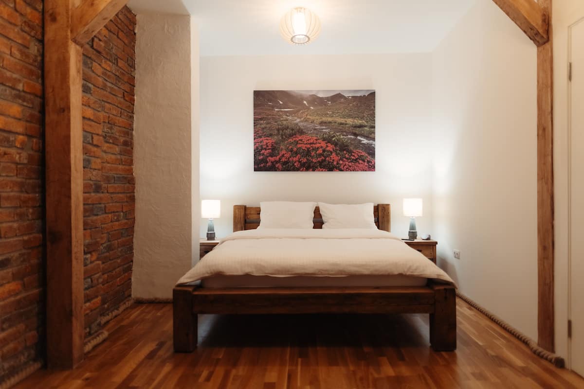 A bedroom with a wooden bed, white bedding, a brick wall, and a painting of a landscape.