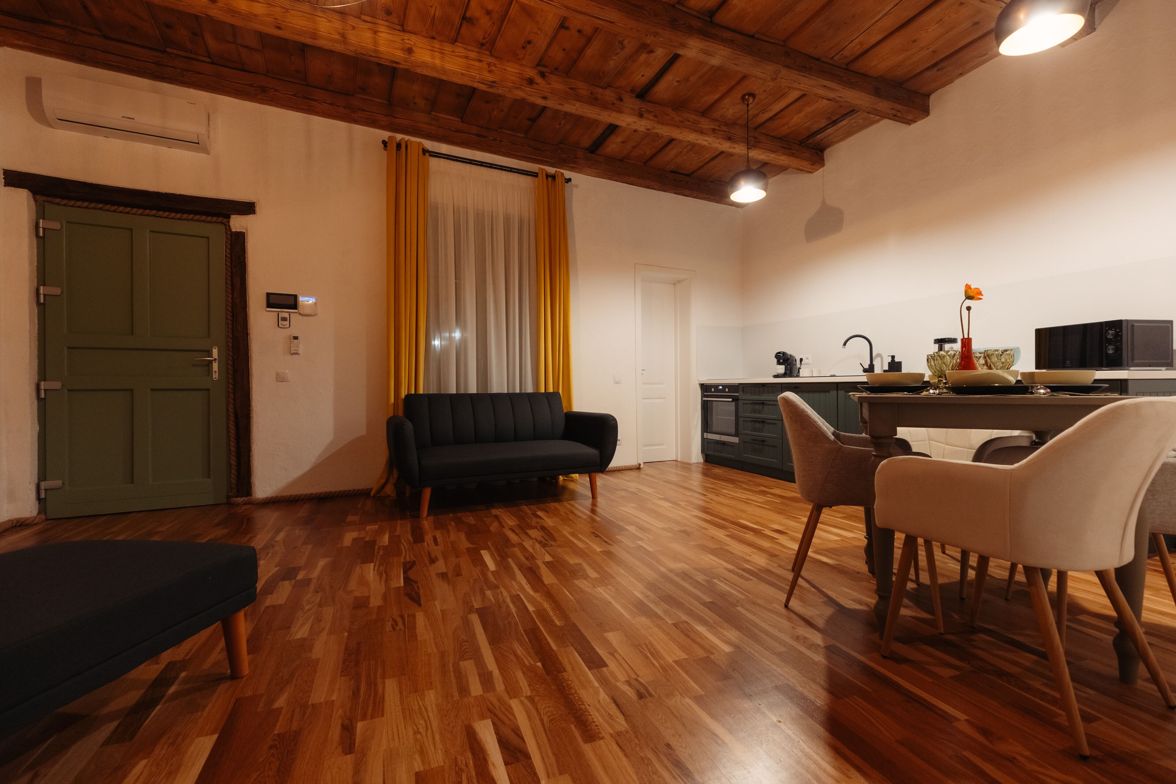 A living room with a sofa, chairs, wooden flooring, a green door, and a kitchen area with a microwave and cabinets.