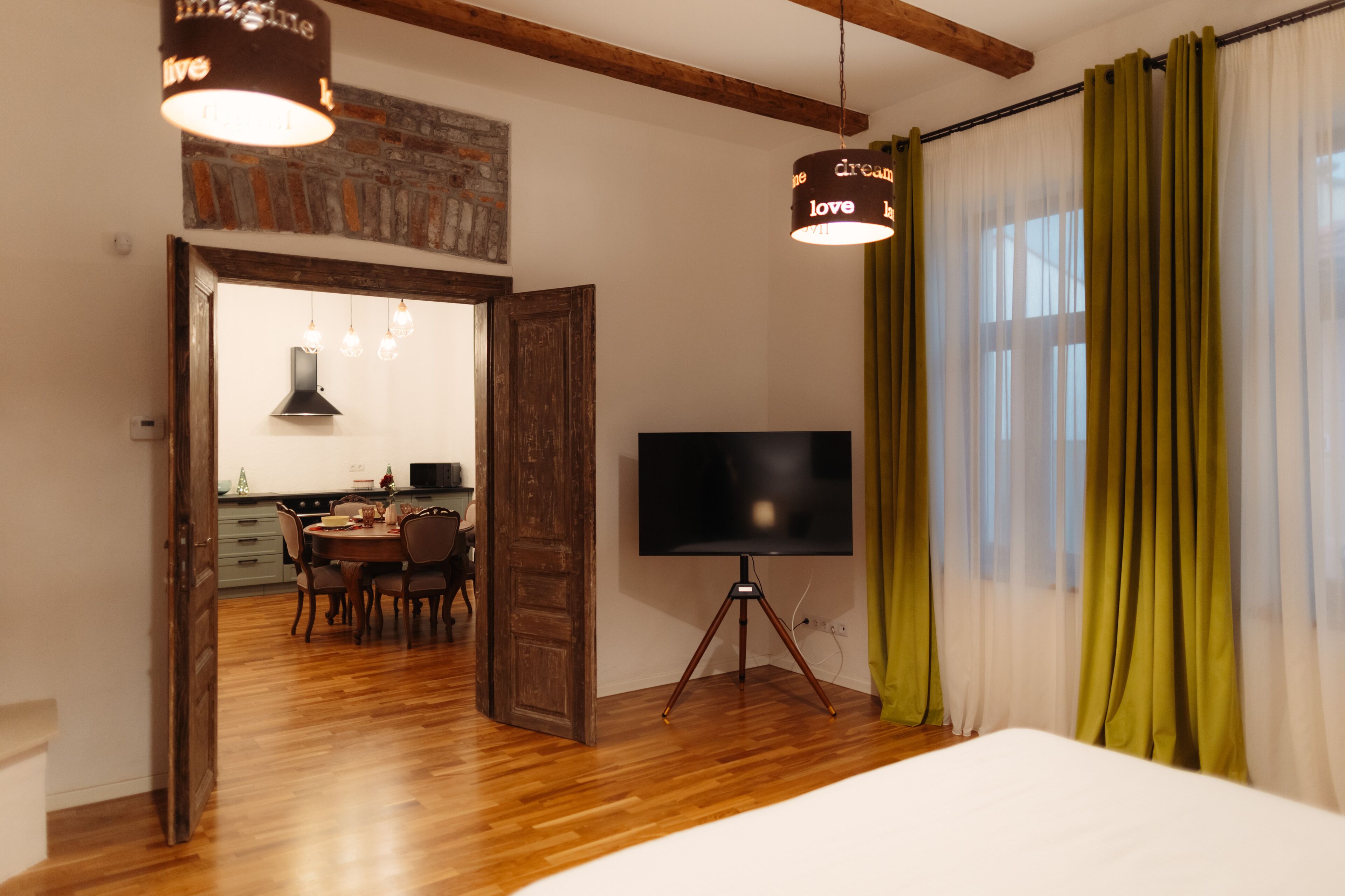 A room with a bed, a TV on a tripod stand, a dining area with chairs and a table, and a kitchen visible through an open door.