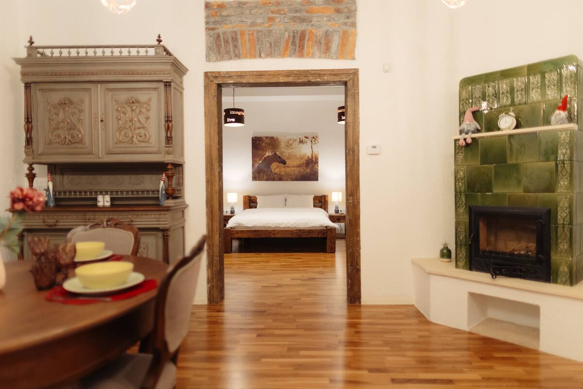 A room with a wooden dining table, a bed with a white bedspread, a fireplace with a green tiled surround, and a wall-mounted shelf with decorative items.
