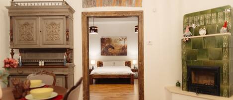 Una habitación con una mesa de comedor de madera, una cama con cubrecama blanco, una chimenea con revestimiento de azulejos verdes y un estante de pared con adornos.
