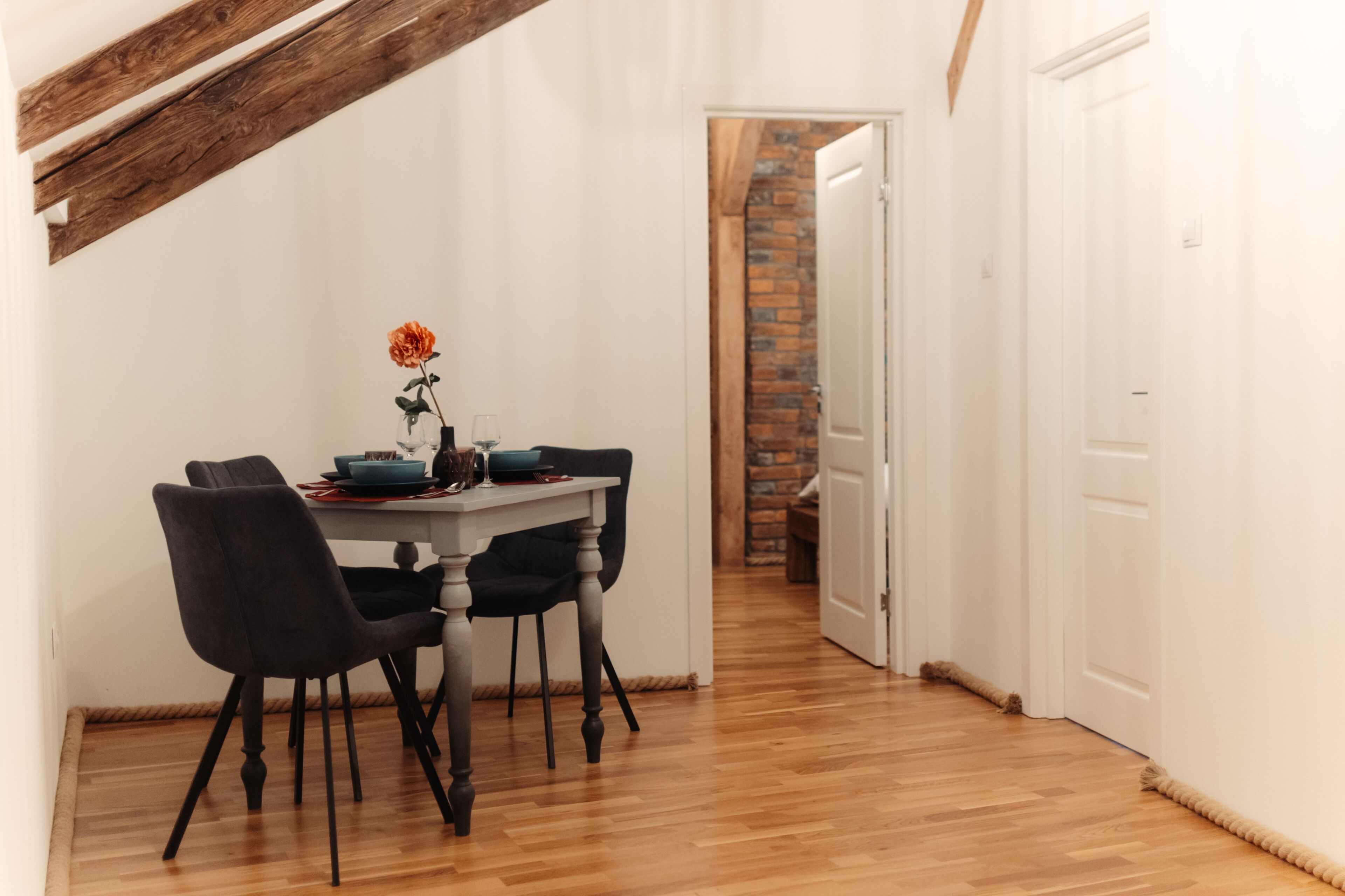 A dining area with a wooden table, black chairs, and a vase with a single flower. A brick wall and a white door are visible in the background.