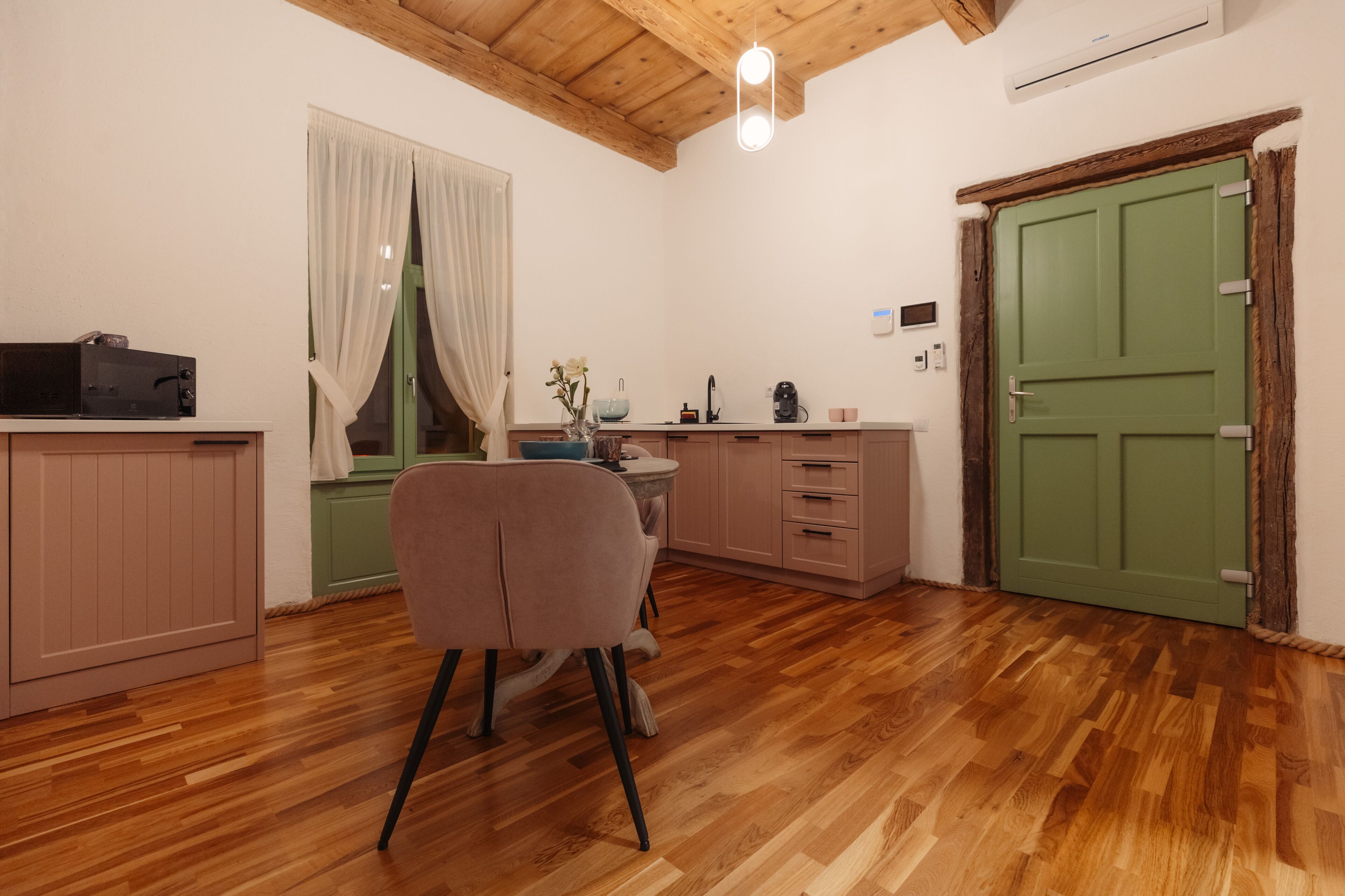 A cozy kitchen with wooden flooring, a dining area with a chair, a window with curtains, and a green door.