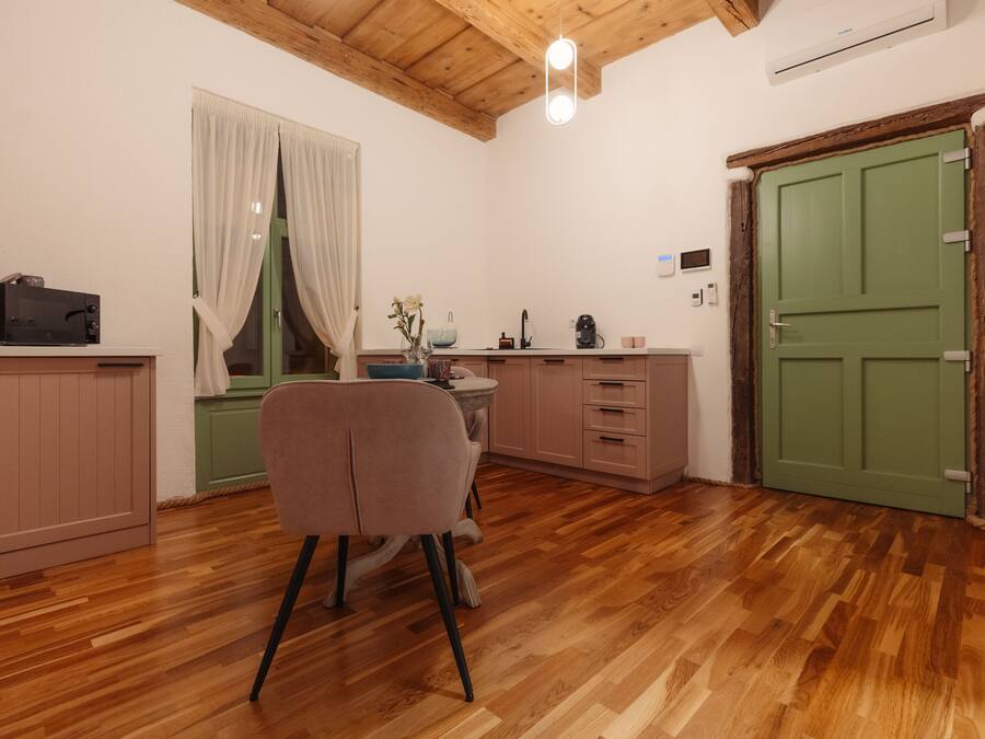 A cozy kitchen with wooden flooring, a dining area with a chair, a window with curtains, and a green door.