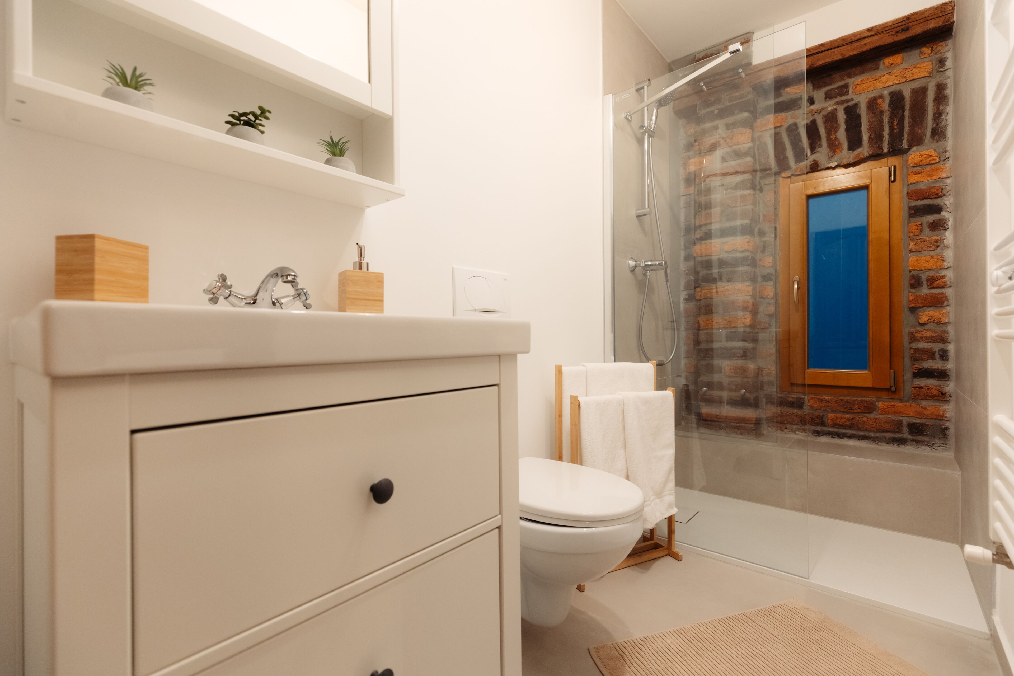 Een badkamer met een moderne wastafel, toilet, douche met glazen deur en een accentmuur van baksteen.