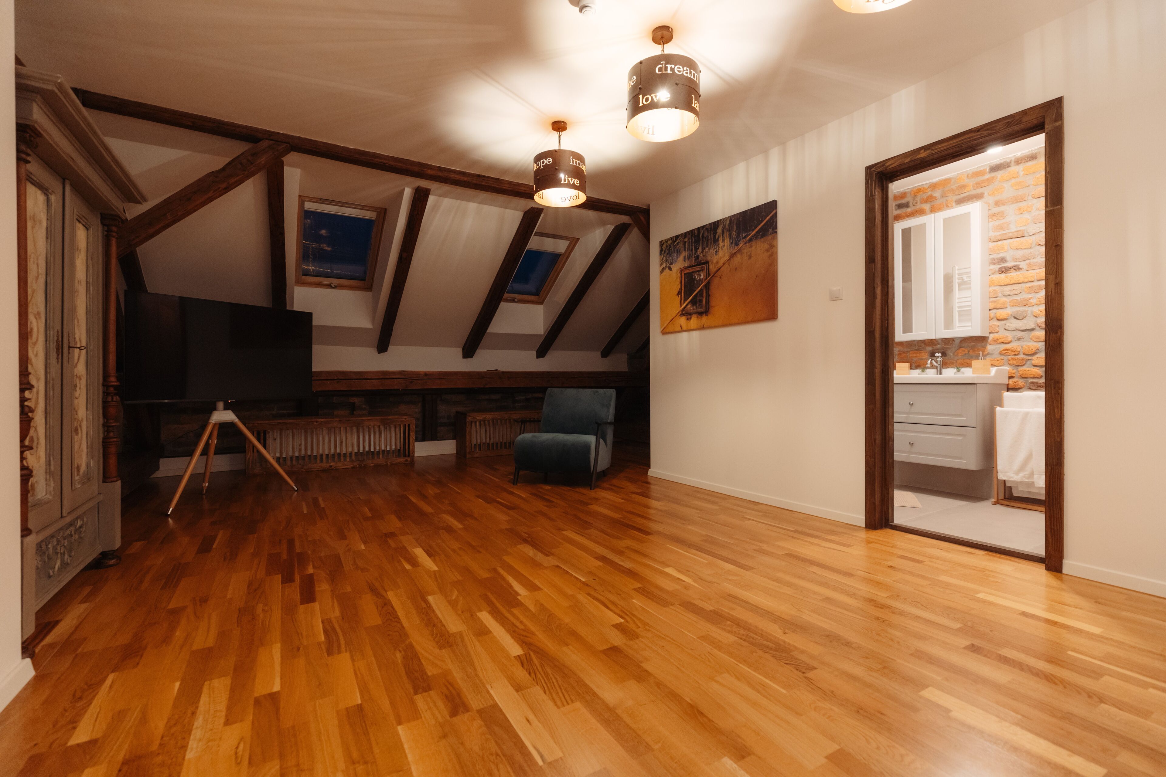 Une chambre spacieuse aménagée en mezzanine, avec un plancher en bois, des poutres apparentes, une grande fenêtre et une vue sur la salle de bain.