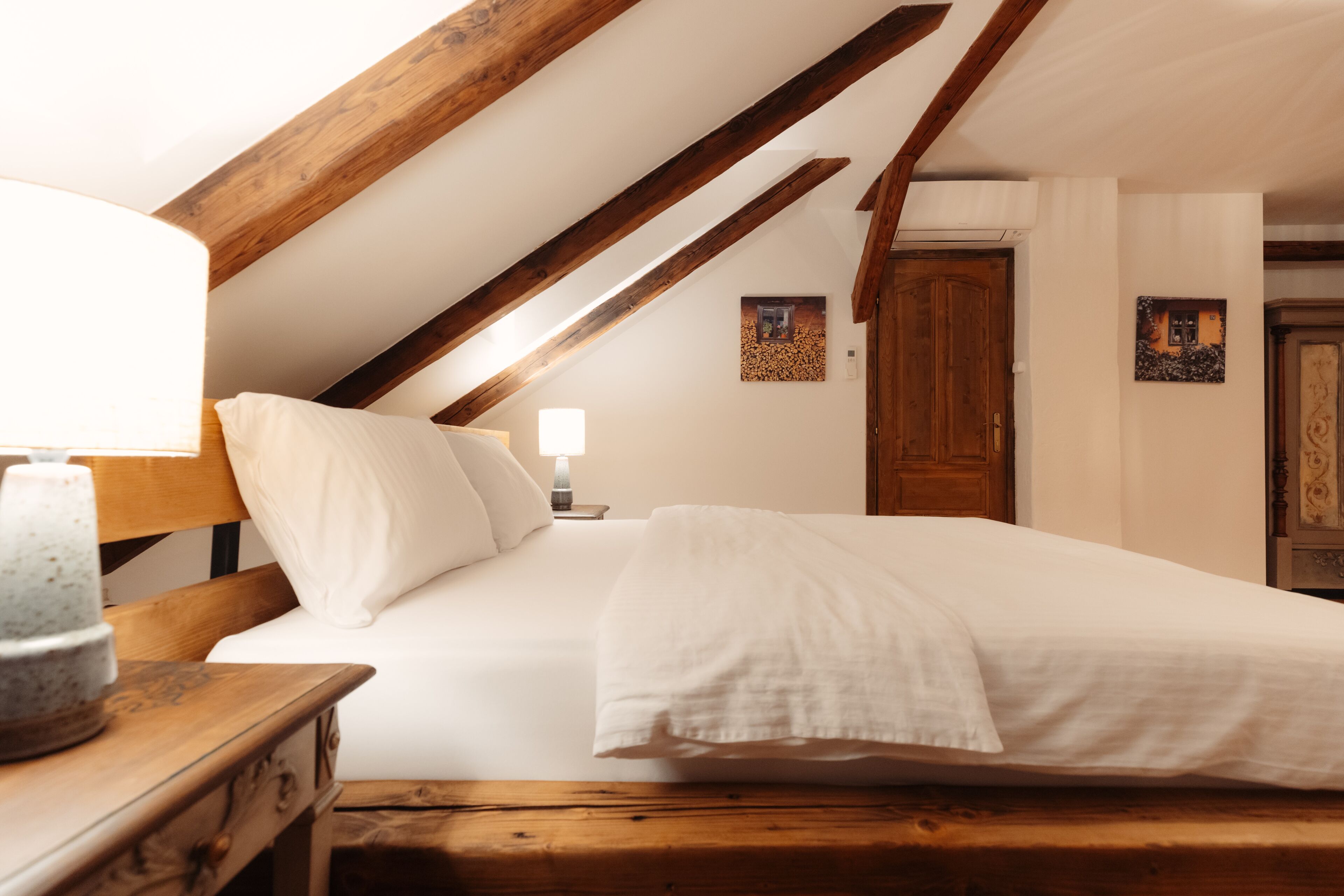 A bedroom with a wooden ceiling, a bed with white linens, a bedside table with a lamp, and a wooden bench.