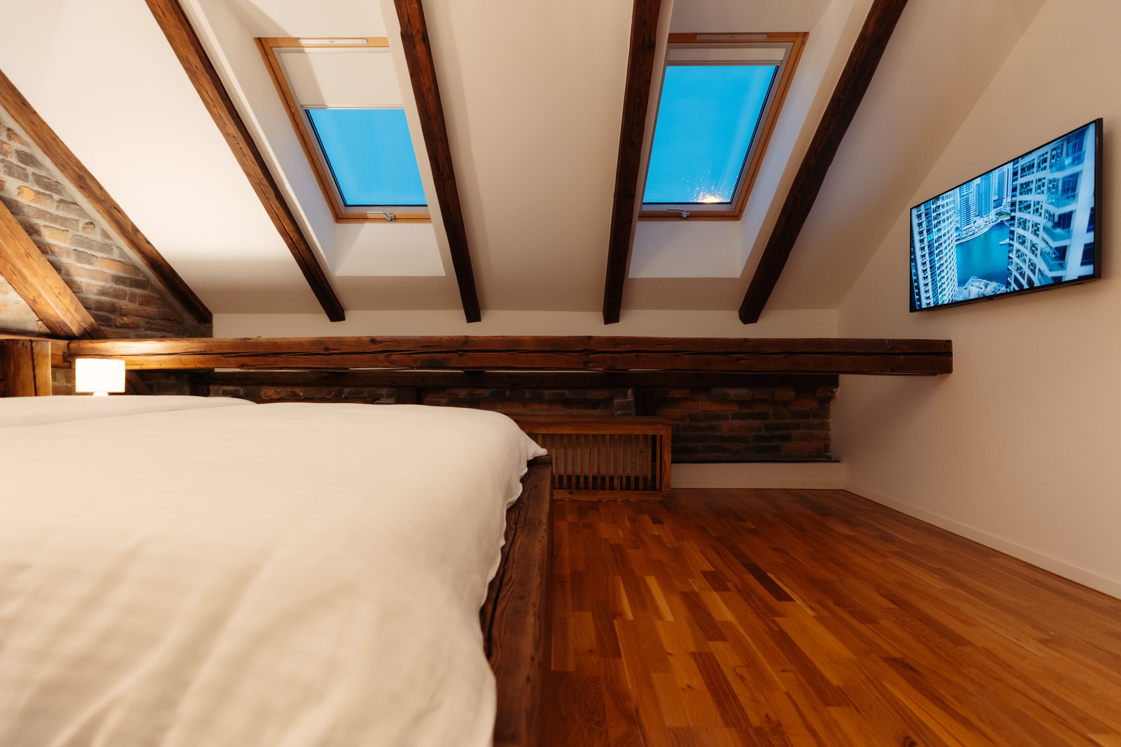 Une chambre avec un plafond en pente, des fenêtres de toit, des poutres en bois, un lit, un banc et un tableau représentant un paysage urbain.