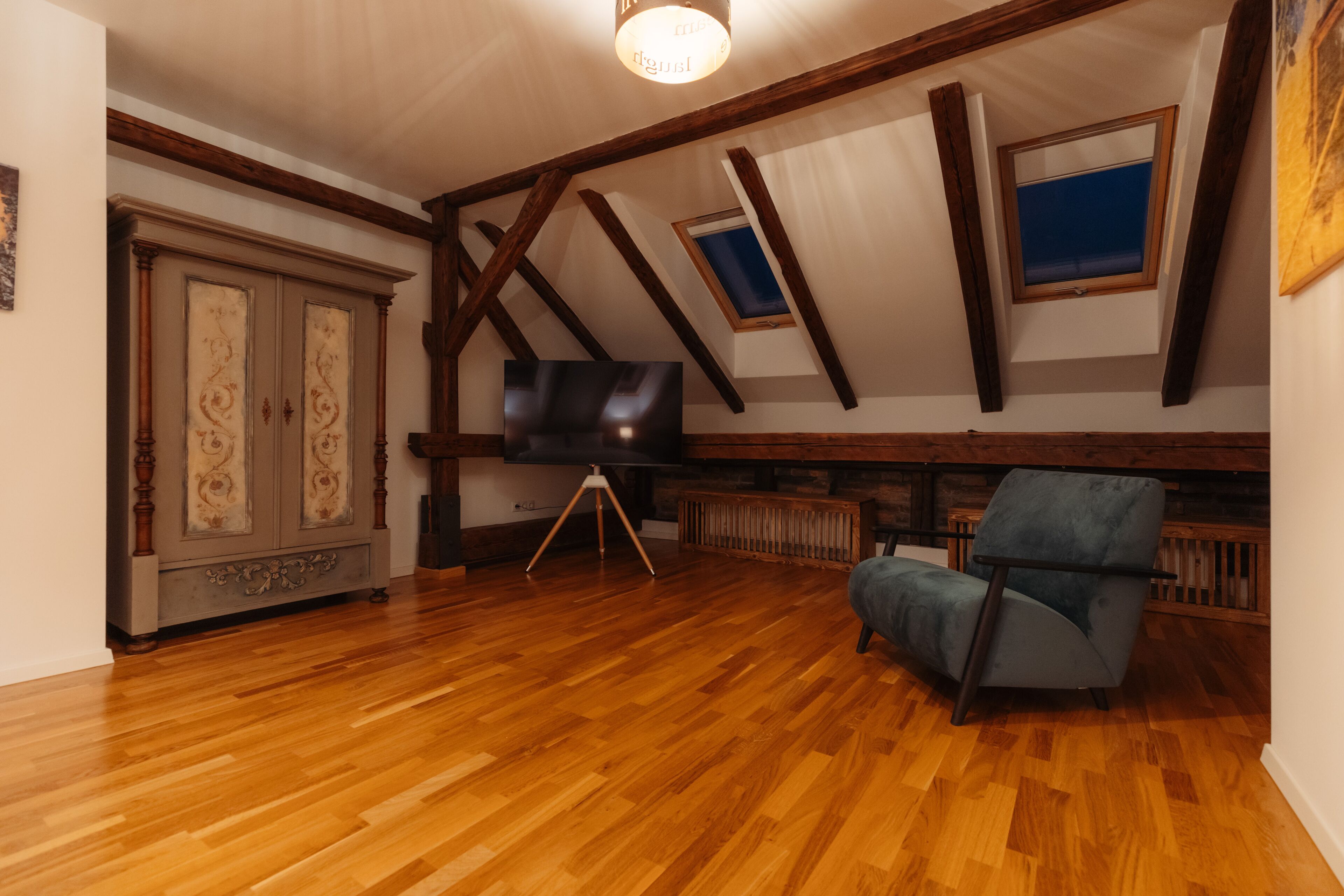 Una habitación con suelo de madera, un televisor sobre un trípode, un sillón azul y un mueble con paneles decorativos.