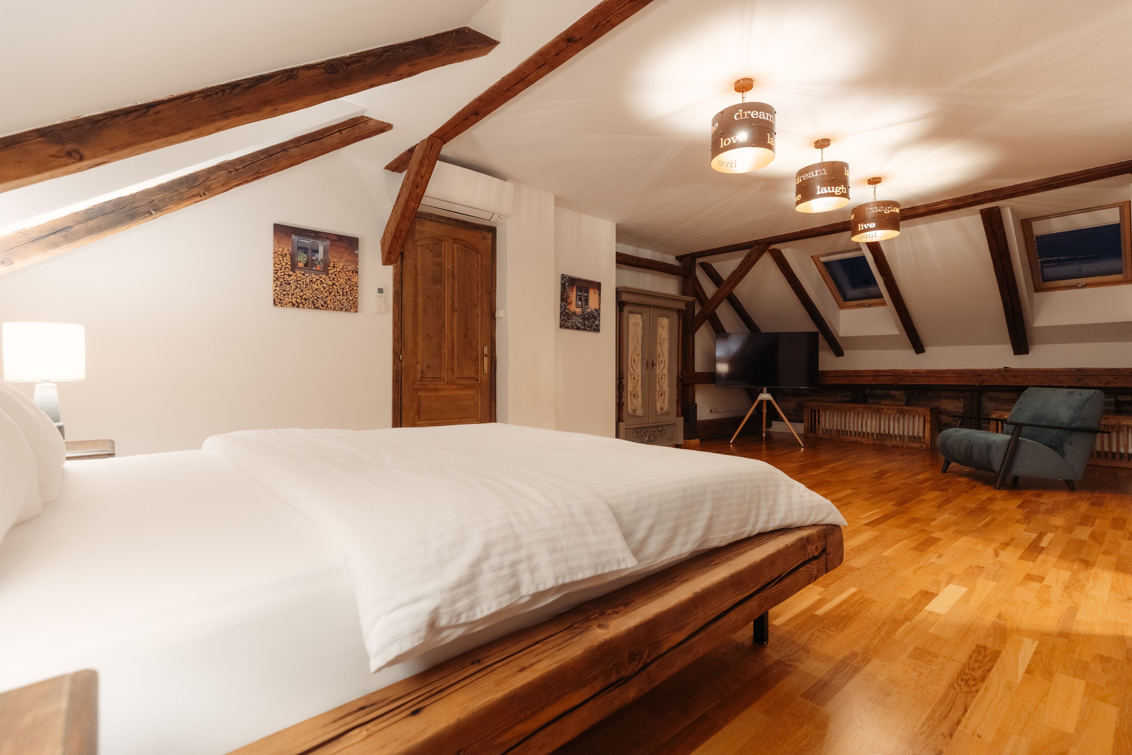 A bedroom with a wooden bed, a chair, and a desk with a chair. There are wooden beams on the ceiling and a picture on the wall.