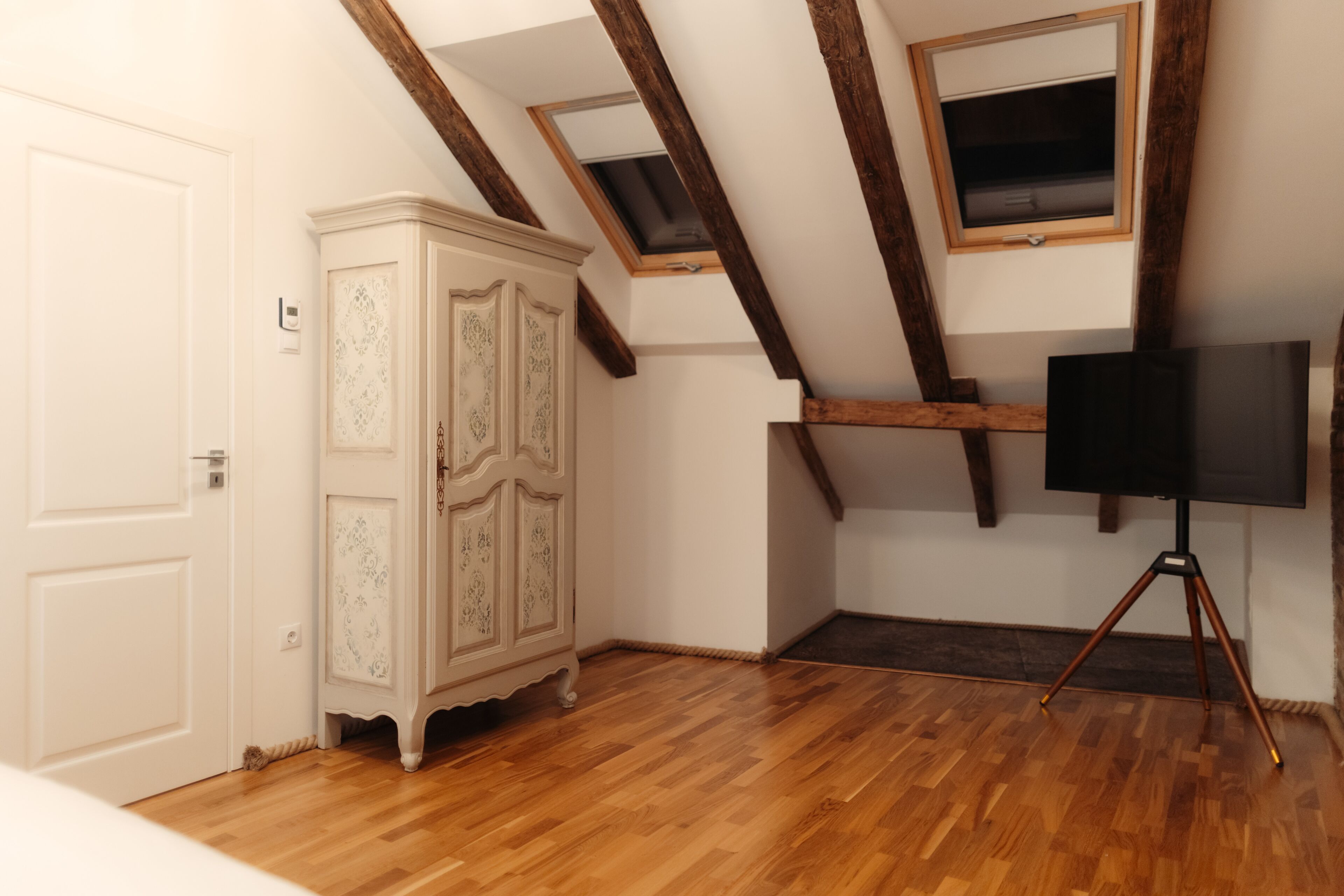 A room with wooden flooring, a white wardrobe, a television on a tripod stand, and a sloped ceiling with wooden beams.