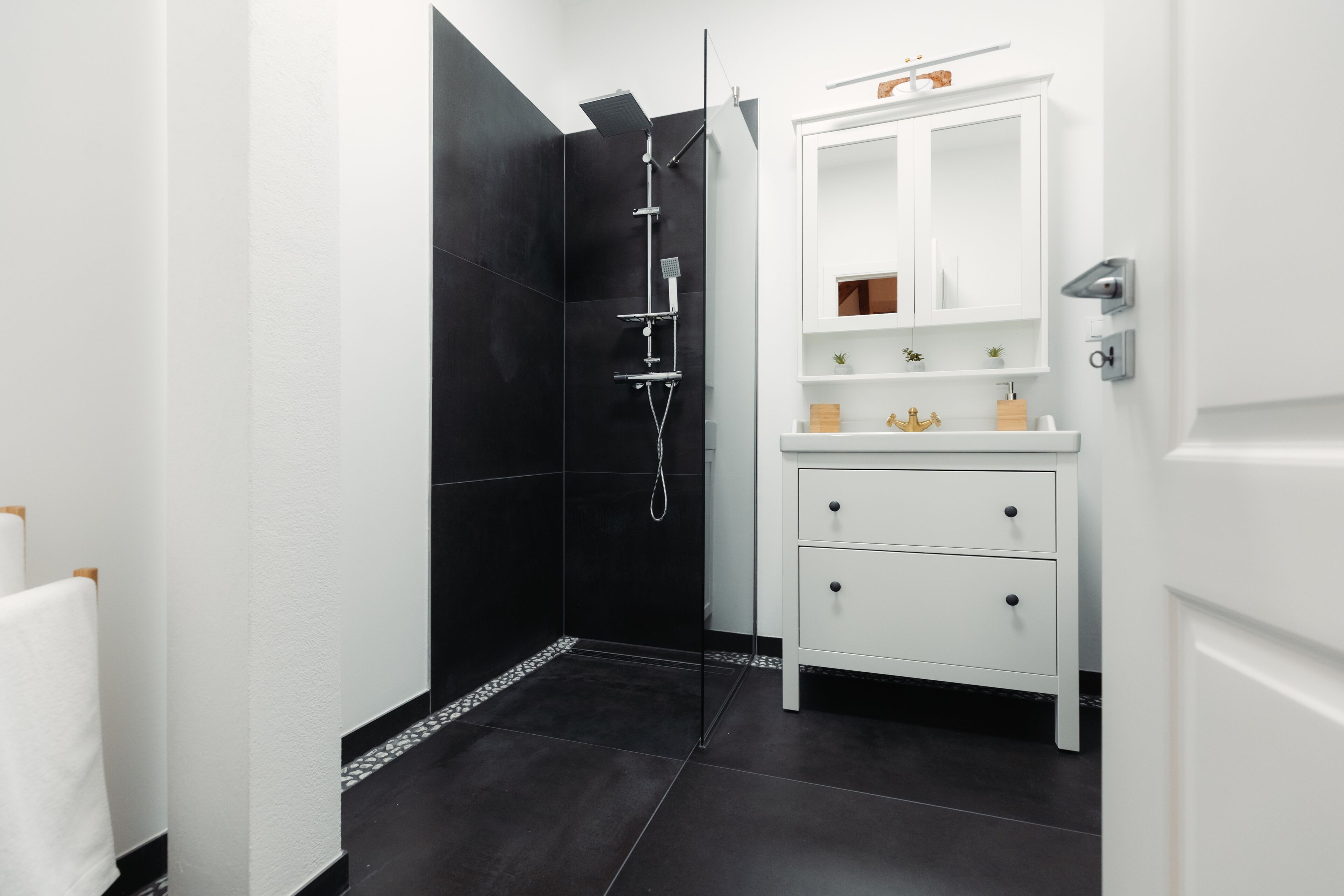 Een moderne badkamer met een douche betegeld in zwart, witte kastjes en een klein plankje met toiletartikelen.