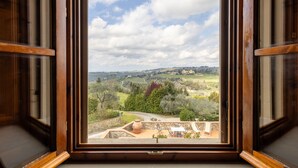 Vista a través de una ventana con marco de madera, que da a un paisaje con colinas onduladas, árboles y un edificio lejano.
