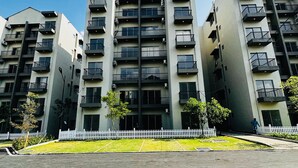 A multi-story residential building with balconies, a white picket fence, and a paved driveway.