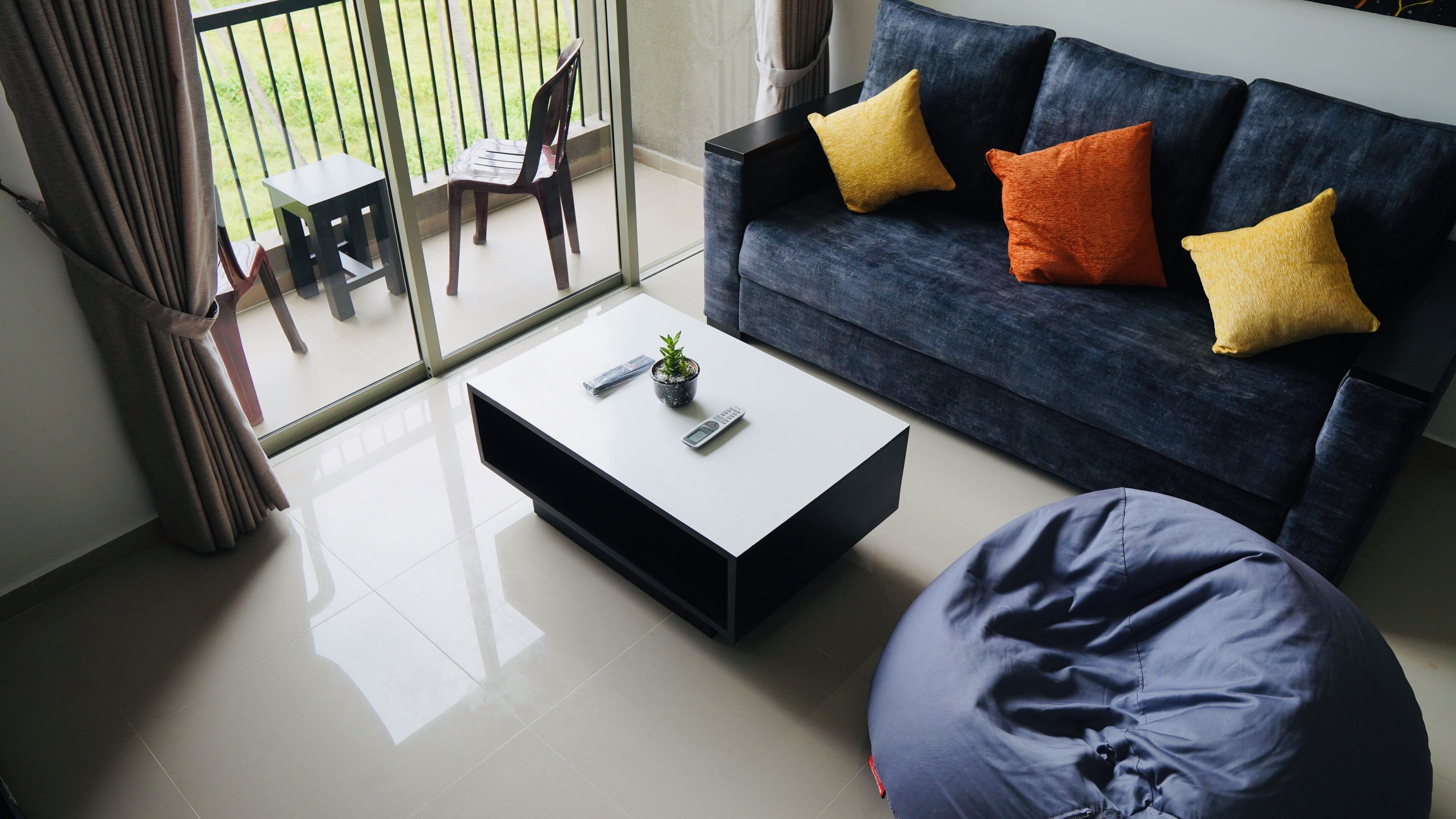Uma sala de estar moderna com um sofá azul escuro, duas almofadas amarelas, uma mesa de centro branca com um controle remoto e uma pequena planta, e uma poltrona de capacho azul.