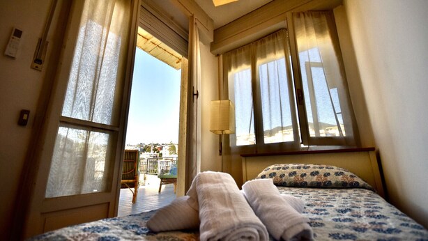 A hotel room with a bed, two rolled towels, a window with curtains, and a lamp.