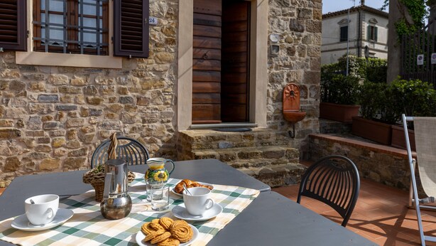Un bâtiment en pierre, une table recouverte d’une nappe à carreaux, une carafe en métal, des tasses et des pâtisseries.