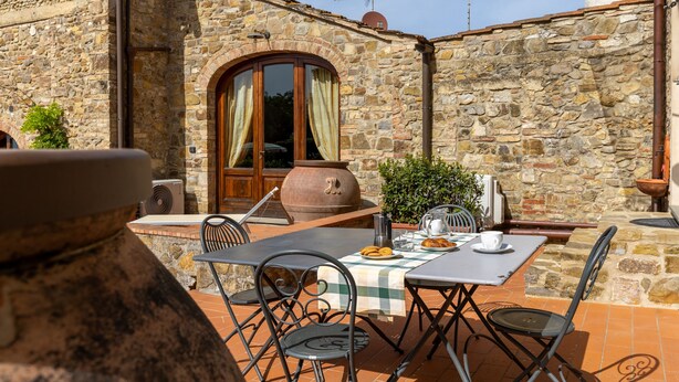 Un edificio de piedra con un patio que incluye una mesa, sillas y una maceta de barro grande.