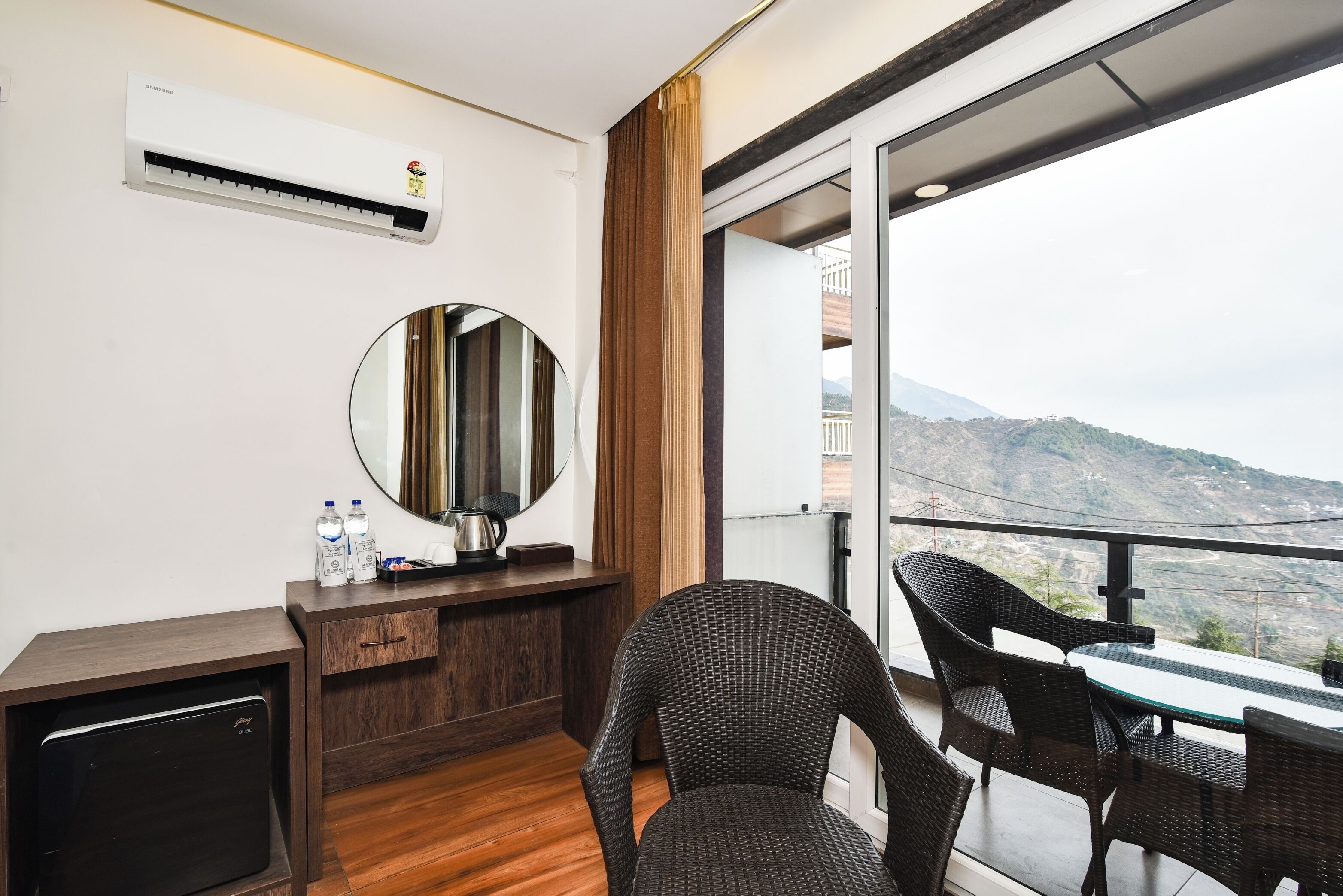 A hotel room with a balcony, a minibar, a round mirror, and a view of a mountainous landscape.