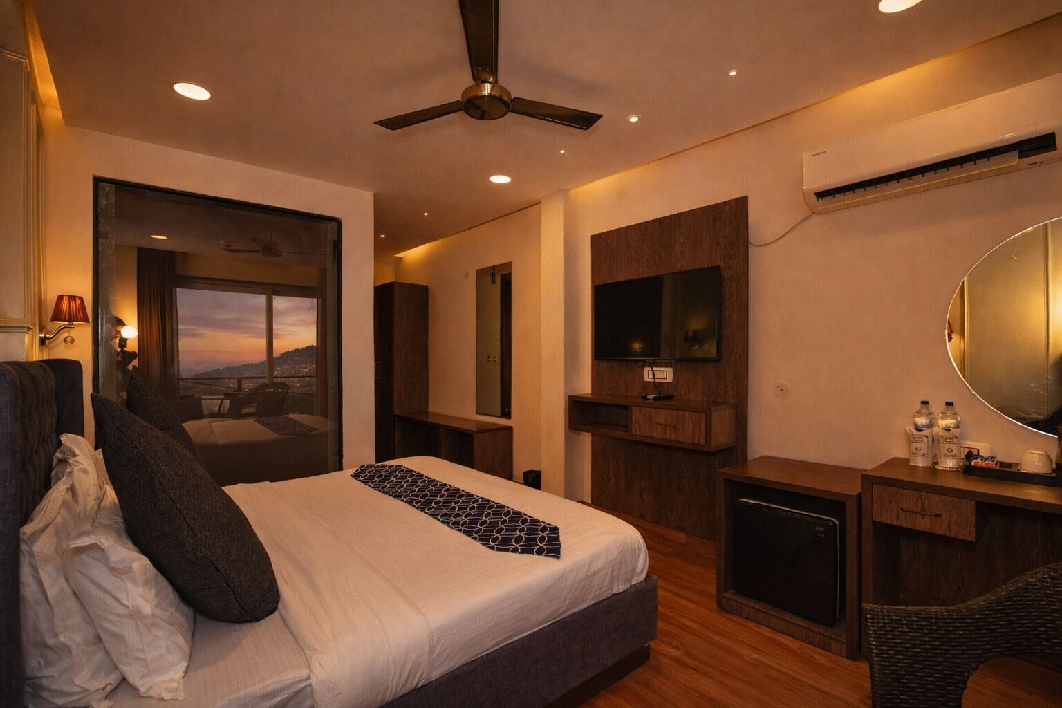 Una habitación de hotel moderna con una cama grande, ventilador de techo, televisor, espejo y vistas a la ciudad.