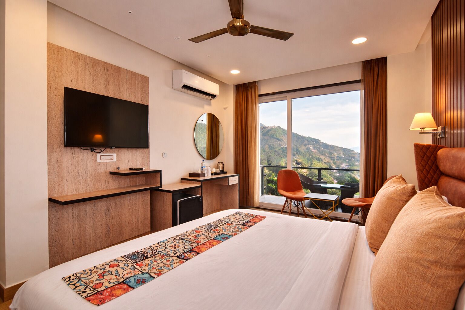 A hotel room with a large bed, a flat-screen TV, a ceiling fan, a balcony with a view of the hills, and a small table with a chair.