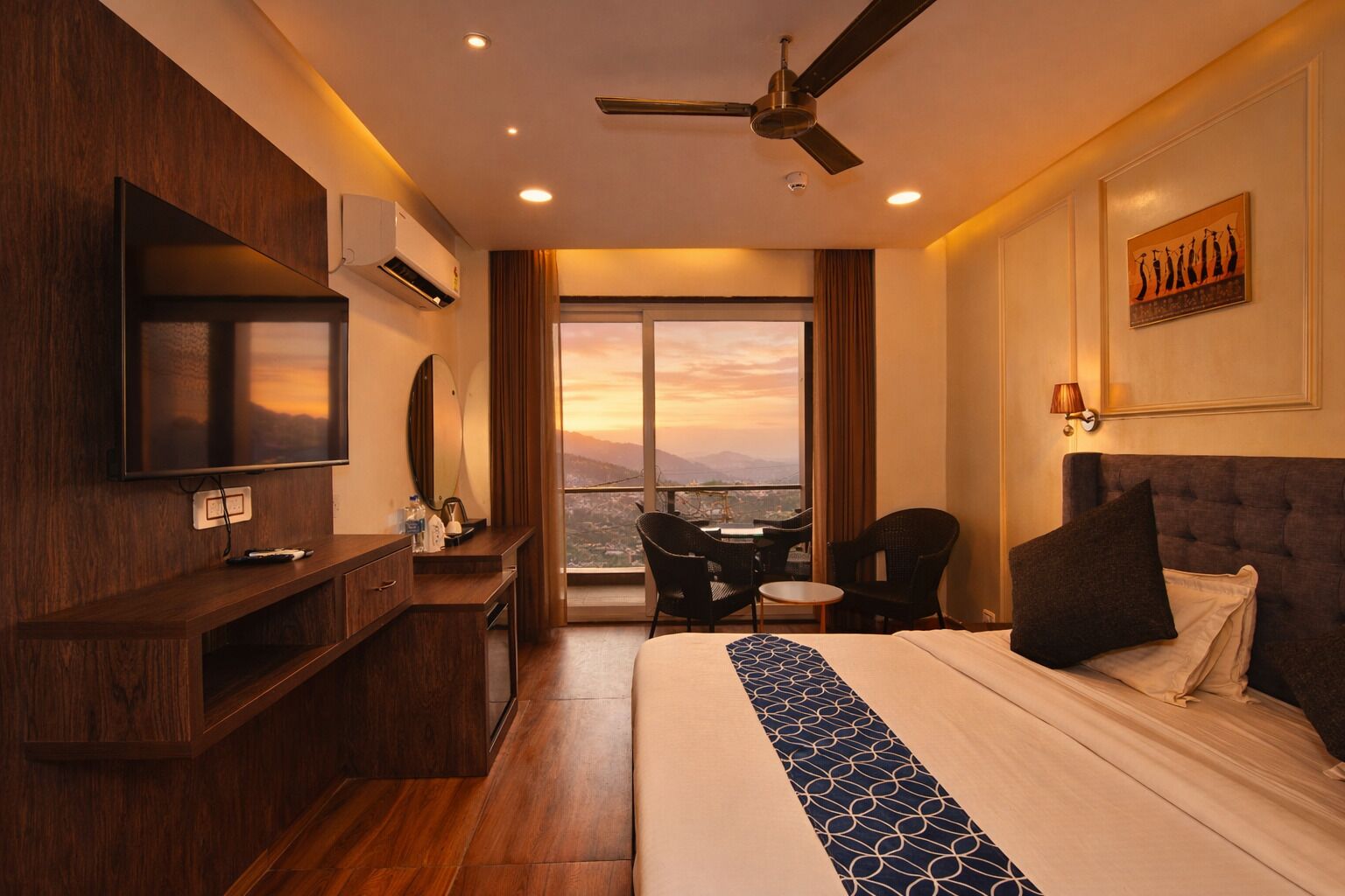 Habitación de hotel con una cama grande, un escritorio con televisor de pantalla plana, ventilador de techo y vista a la ciudad al atardecer.