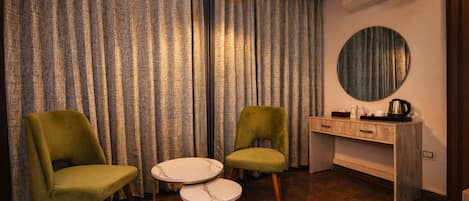 Una habitación con dos sillones verdes, una mesita blanca, un escritorio de madera con un espejo y un equipo de aire acondicionado.