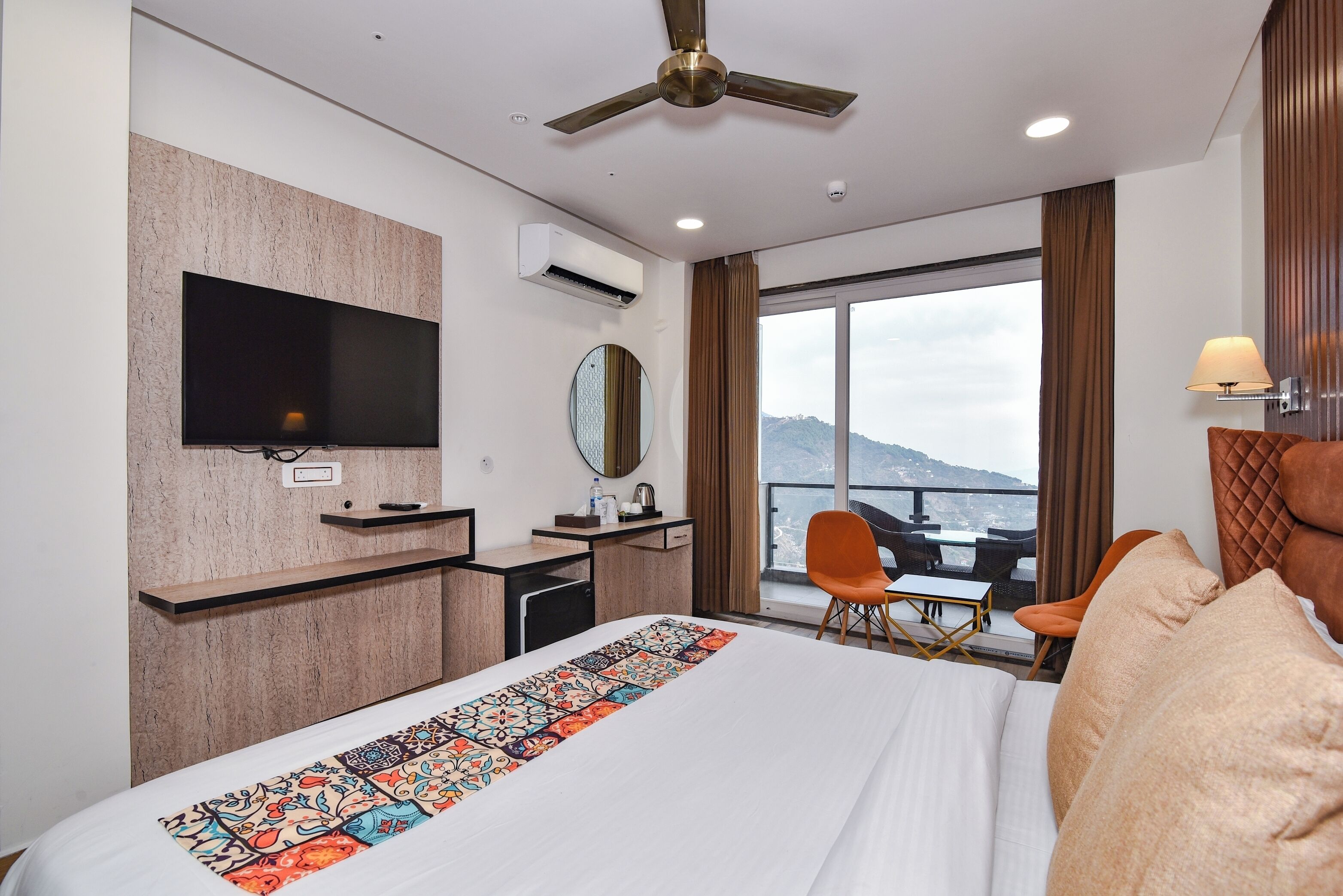 Habitación de hotel con cama, escritorio, televisión, espejo, ventana con vistas y ventilador de techo.