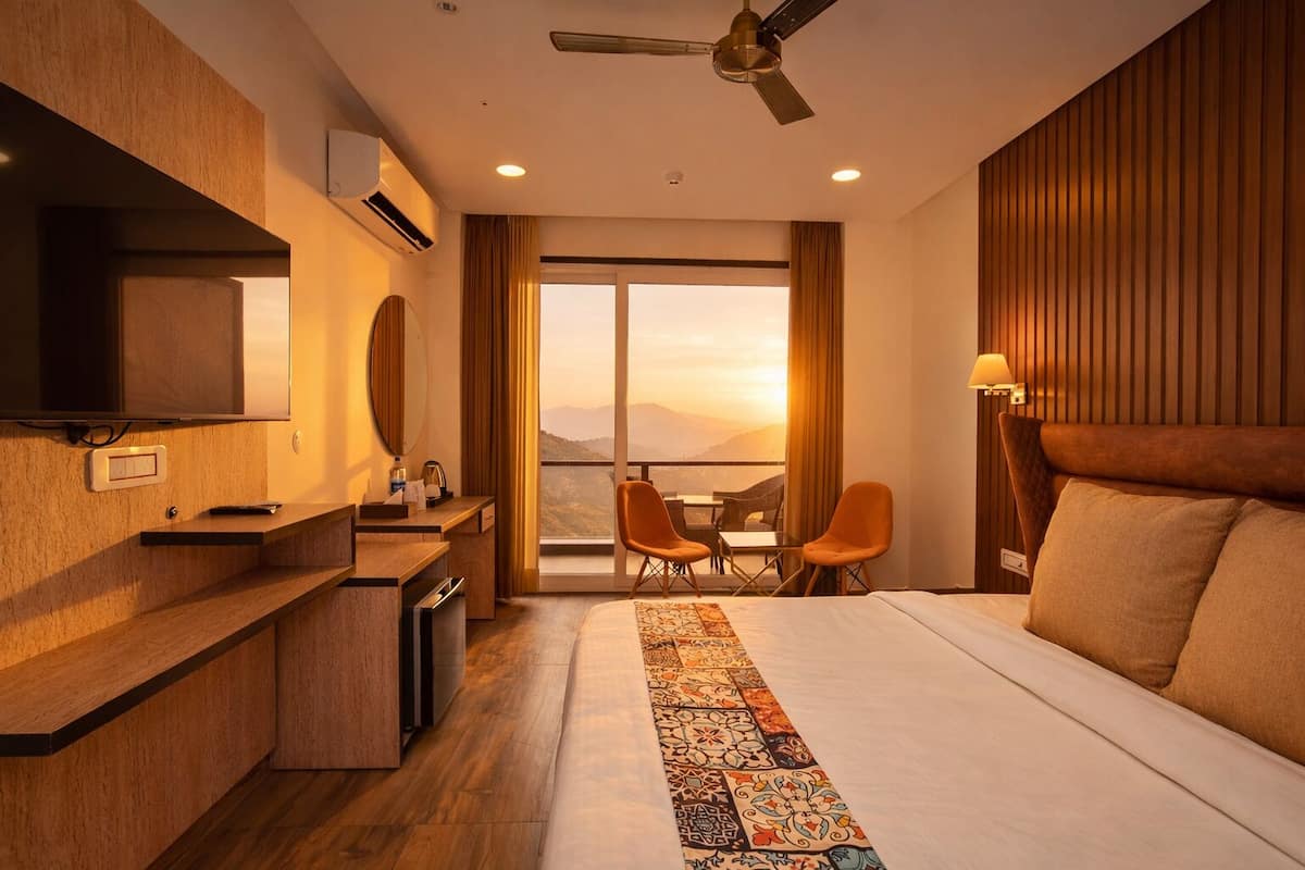 A hotel room with a bed, a desk with a TV, a chair, a table, and a view of the mountains.