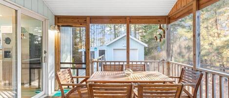 A covered outdoor deck with a wooden table and chairs, large windows, and a view of a house in the background.