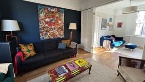 A living room with a dark blue wall, a colorful abstract painting, a black sofa with patterned cushions, a coffee table with books and decorative items, and a bright window with natural light. - Dreamy 3BD House W/large Patio - Stoke Newington! (London)