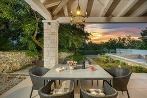 Um pátio externo com um pilar de pedra, cadeiras de vime, uma mesa com uma garrafa e copos, e vista para o pôr do sol.