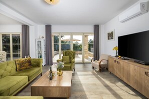 A modern living room with a sofa, armchair, coffee table, TV, and a view through large glass doors.