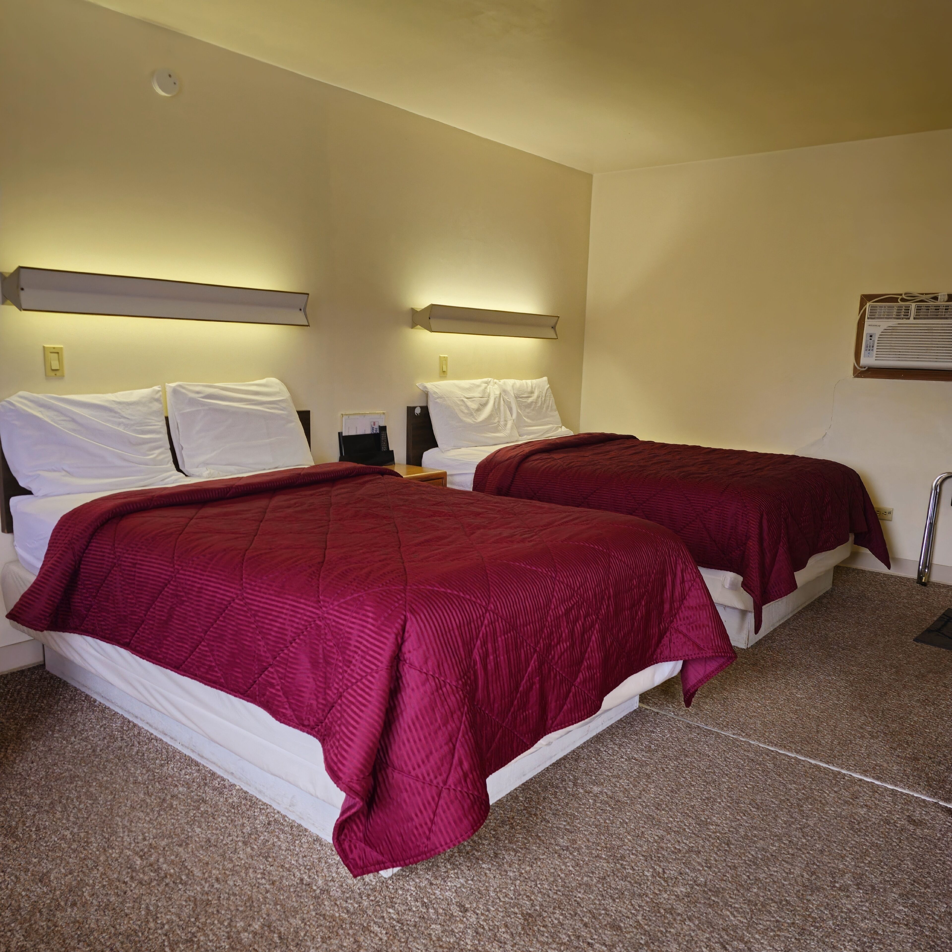 A hotel room with two beds, each with white bedding and a red blanket. There is a nightstand with a lamp and a remote control. The room has beige walls and a ceiling light fixture.