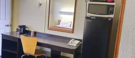 A hotel room with a desk, chair, TV, microwave, and refrigerator.