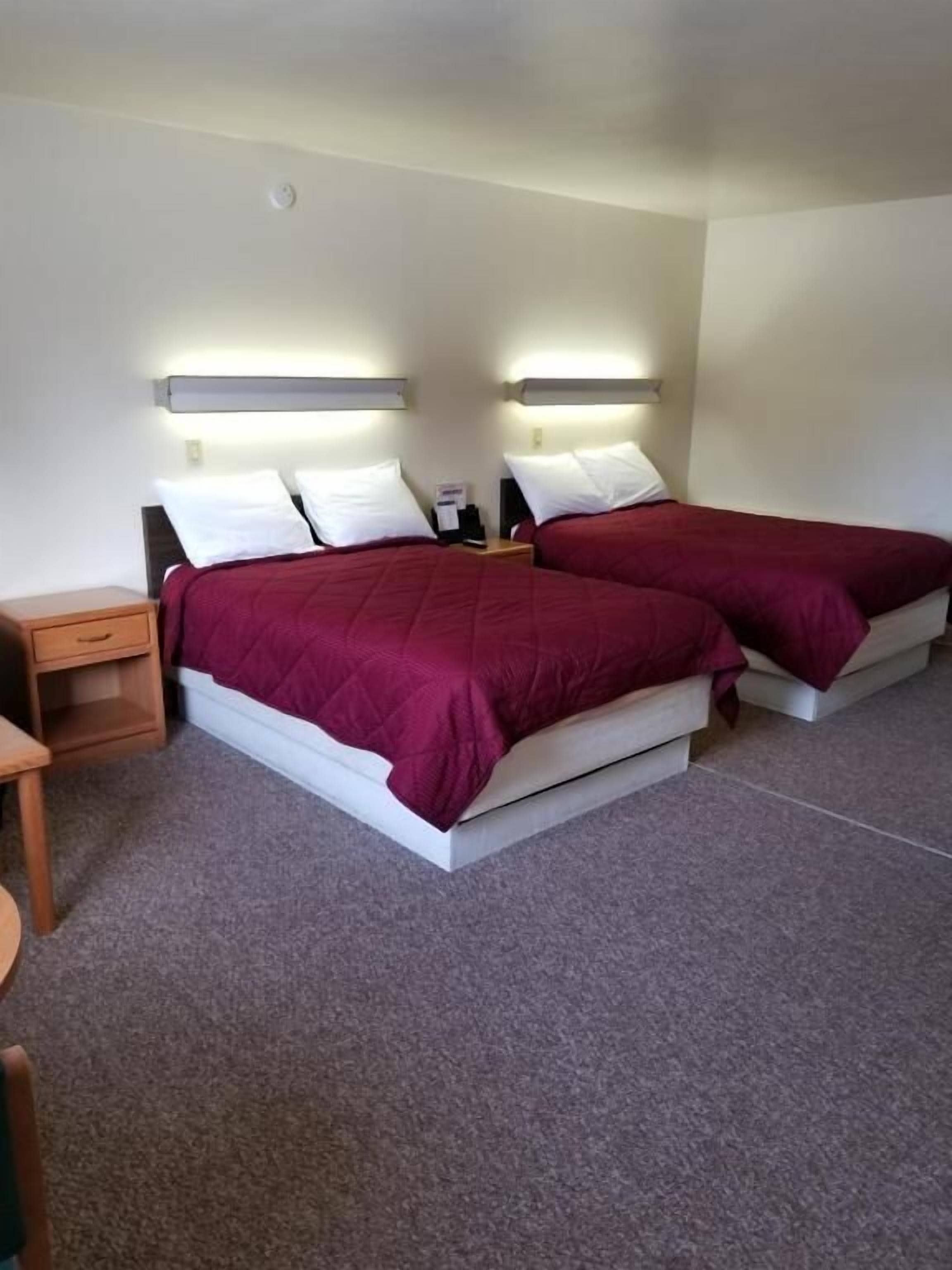 Een hotelkamer met twee bedden, elk met een witte hoofdbord en rode beddengoed. Er is een klein houten nachtkastje met een lamp en een kleine tafel met een stoel.