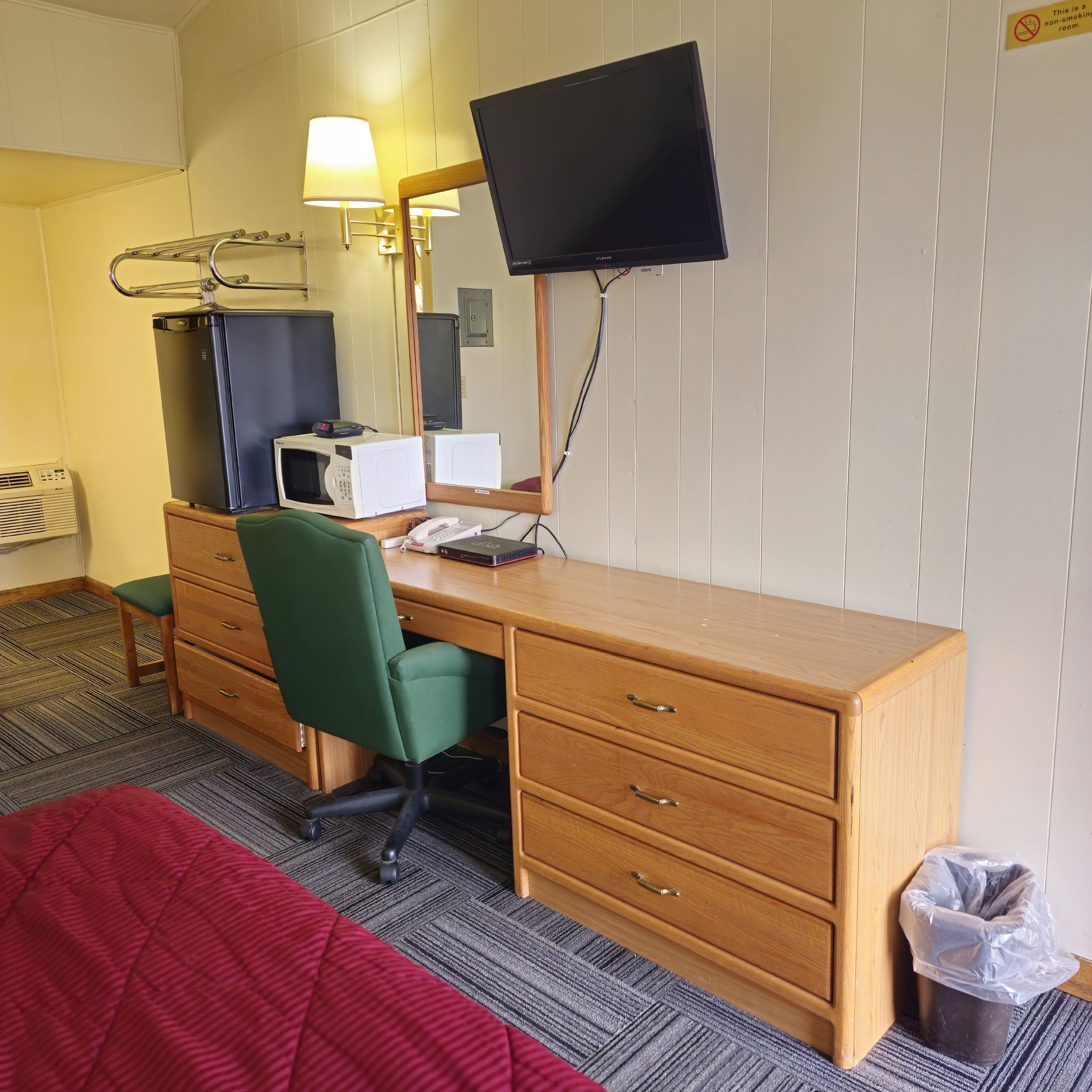 A hotel room with a wooden desk, a green chair, a TV, a microwave, a telephone, a trash bin, and a bed with a red blanket.